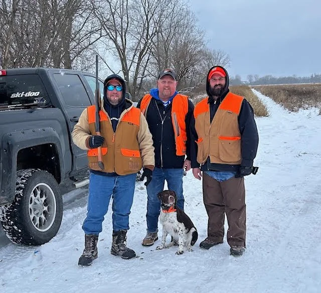 Three men in orange and tan hunting vests standing on a snowy road with a hunting dog and an SUV behind them.
