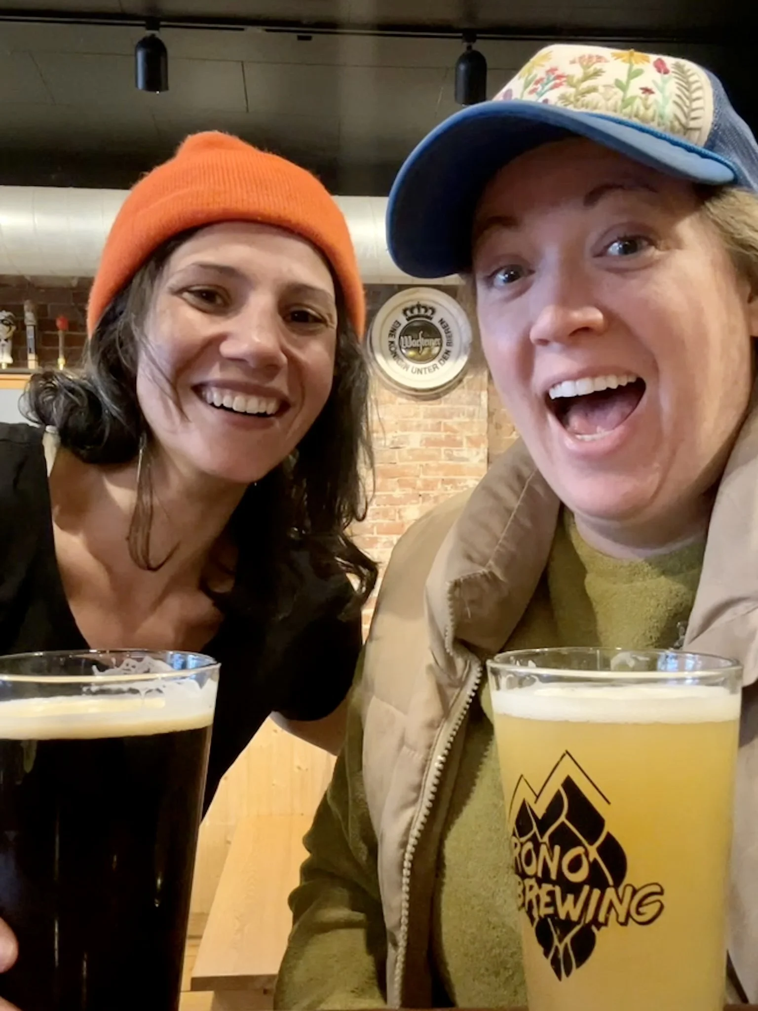 Two women are smiling and enjoying drinks at a brewery or pub, with one woman wearing an orange beanie and the other wearing a cap with a floral design. They are sitting close together with glasses of dark and light beer in front of them.