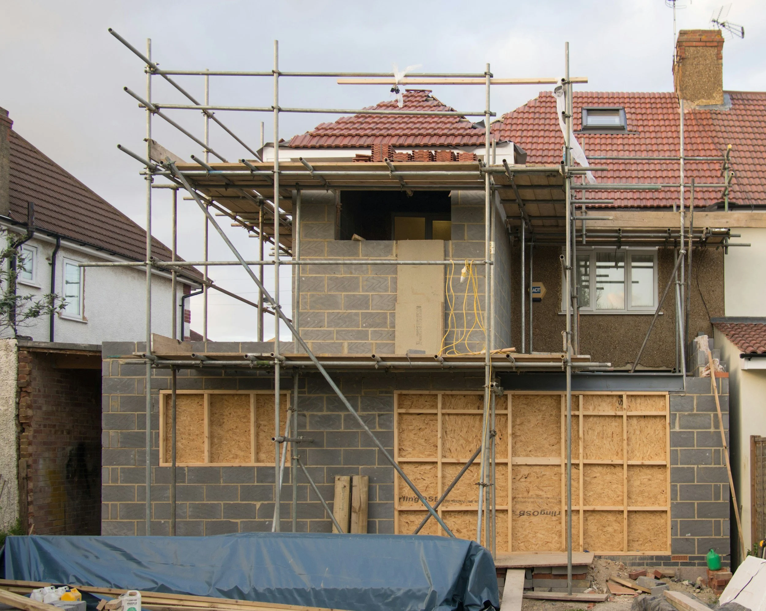 Construction of a house with scaffolding around it, with some brickwork and framing in progress, and a partially completed upper level.