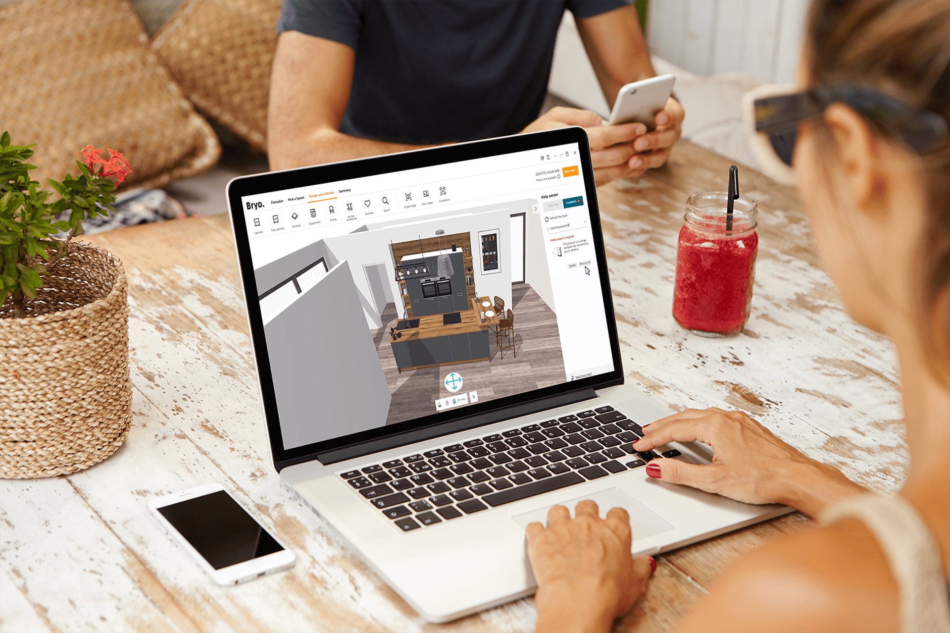 A person using a laptop to view a 3D interior design of a kitchen, with two other people at the table, one holding a smartphone and a red drink with a straw nearby.