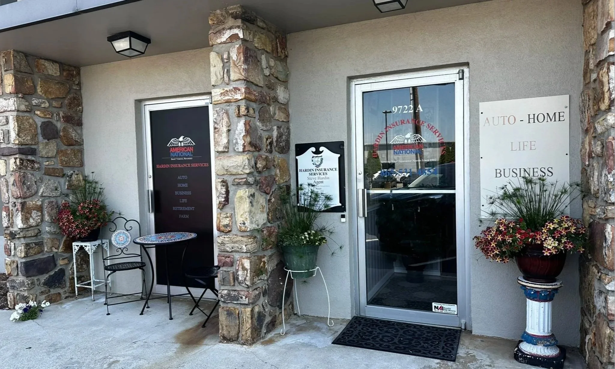 Entrance to an insurance business with glass doors, two signs, potted plants, a small outdoor seating area, and flower arrangements.