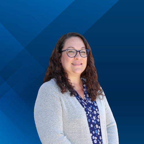 A woman with curly brown hair and glasses smiling, standing in front of a blue geometric background.