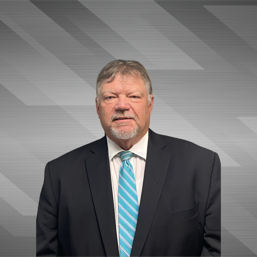 A middle-aged man with greying hair and a beard wearing a black suit, white shirt, and a blue striped tie standing against a grey background with diagonal lines.
