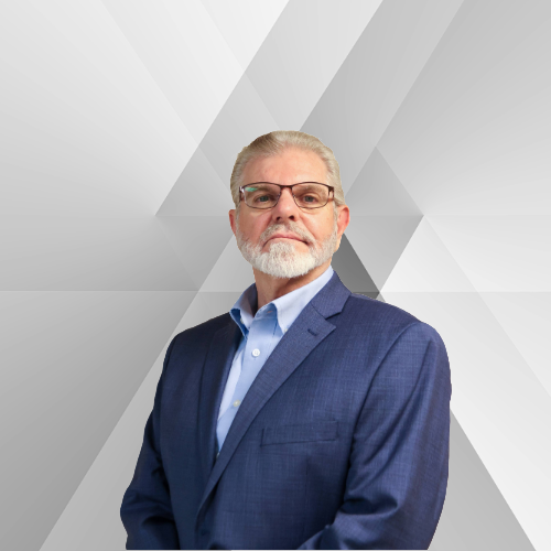 An older man with gray hair, glasses, and a beard wearing a blue suit and light blue shirt against a geometric gray background.