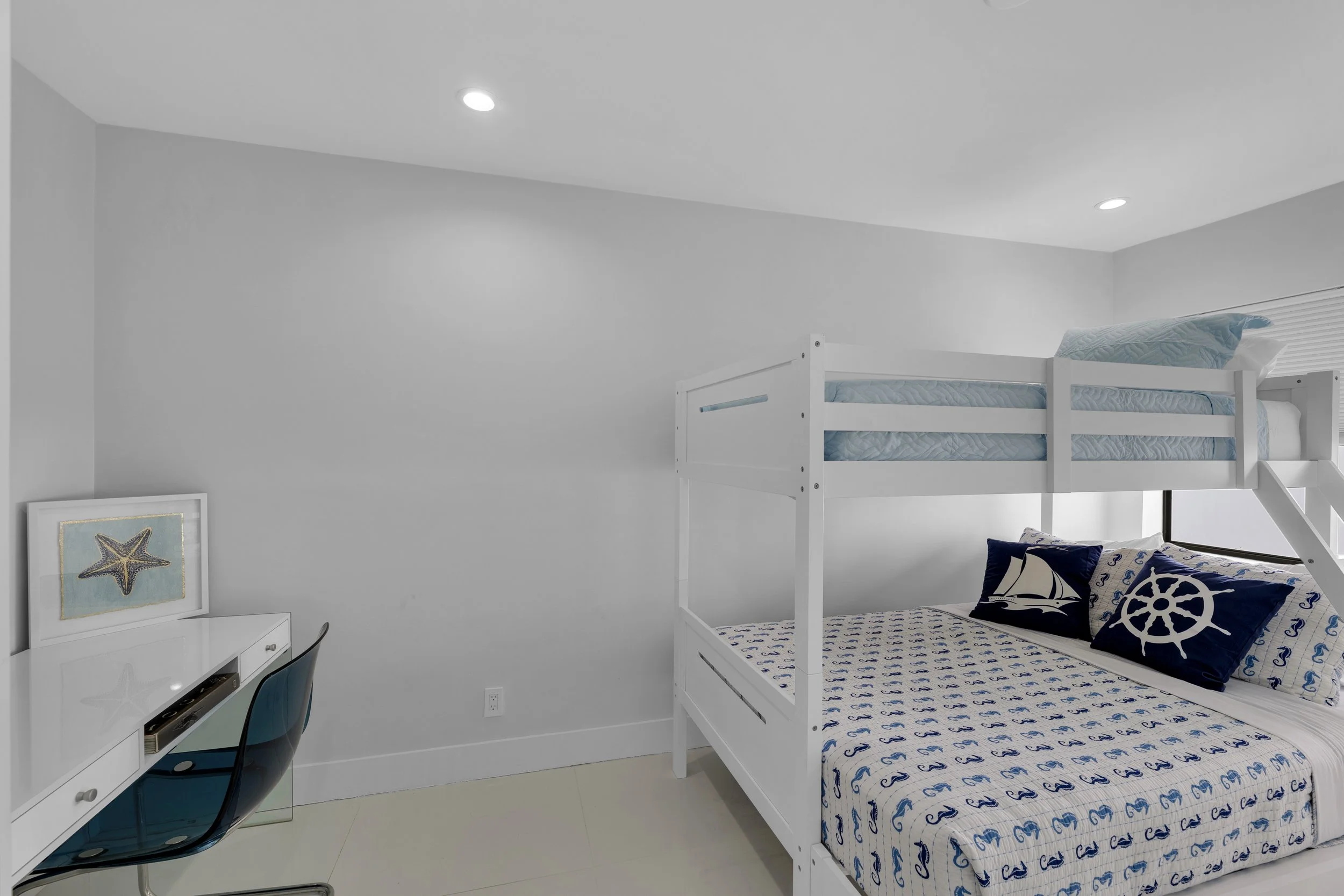 A bedroom with a white bunk bed and nautical-themed bedding, a white desk with a starfish picture on top, and a blue chair, all in a minimalist, bright room.