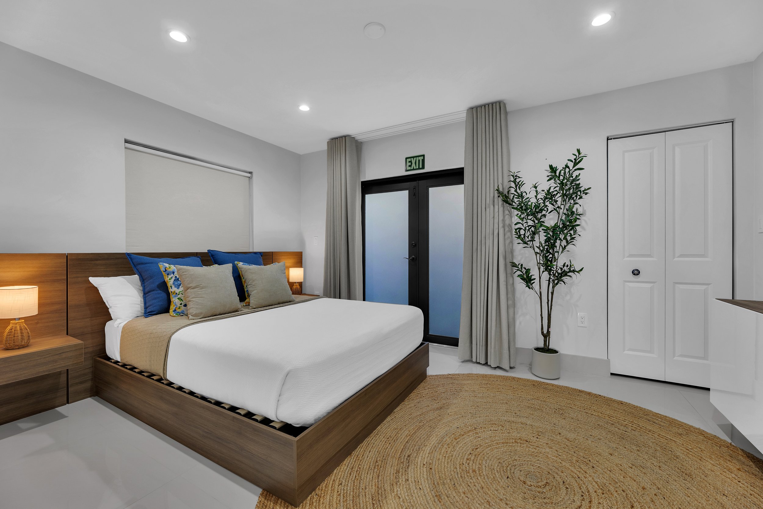 A modern bedroom with a wooden bed frame, white bedding, blue and beige pillows, side tables with lamps, a potted plant near a white closet, gray curtains, and a glass door with an exit sign above.