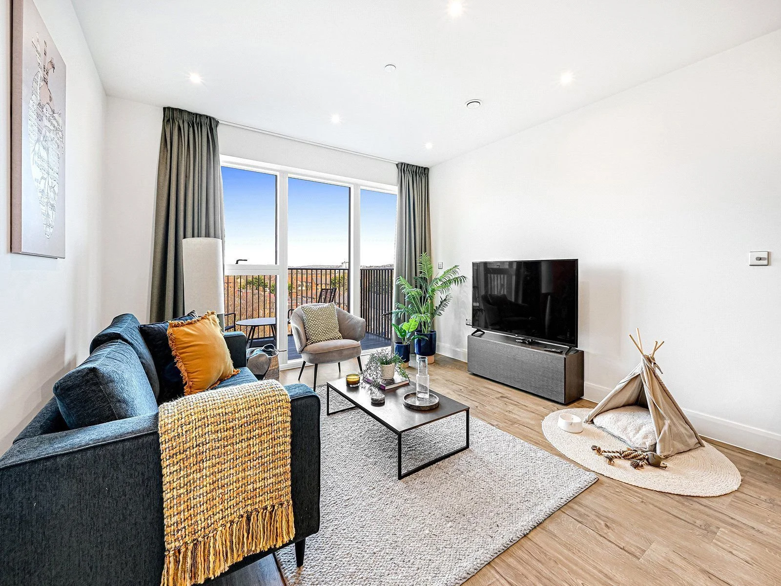 Bright living room with large windows, gray curtains, a dark sofa with yellow pillows, a beige armchair, a coffee table, a flat-screen TV, a potted plant, and a small pet tent with toys on a rug. Located in Fulham Broadway.