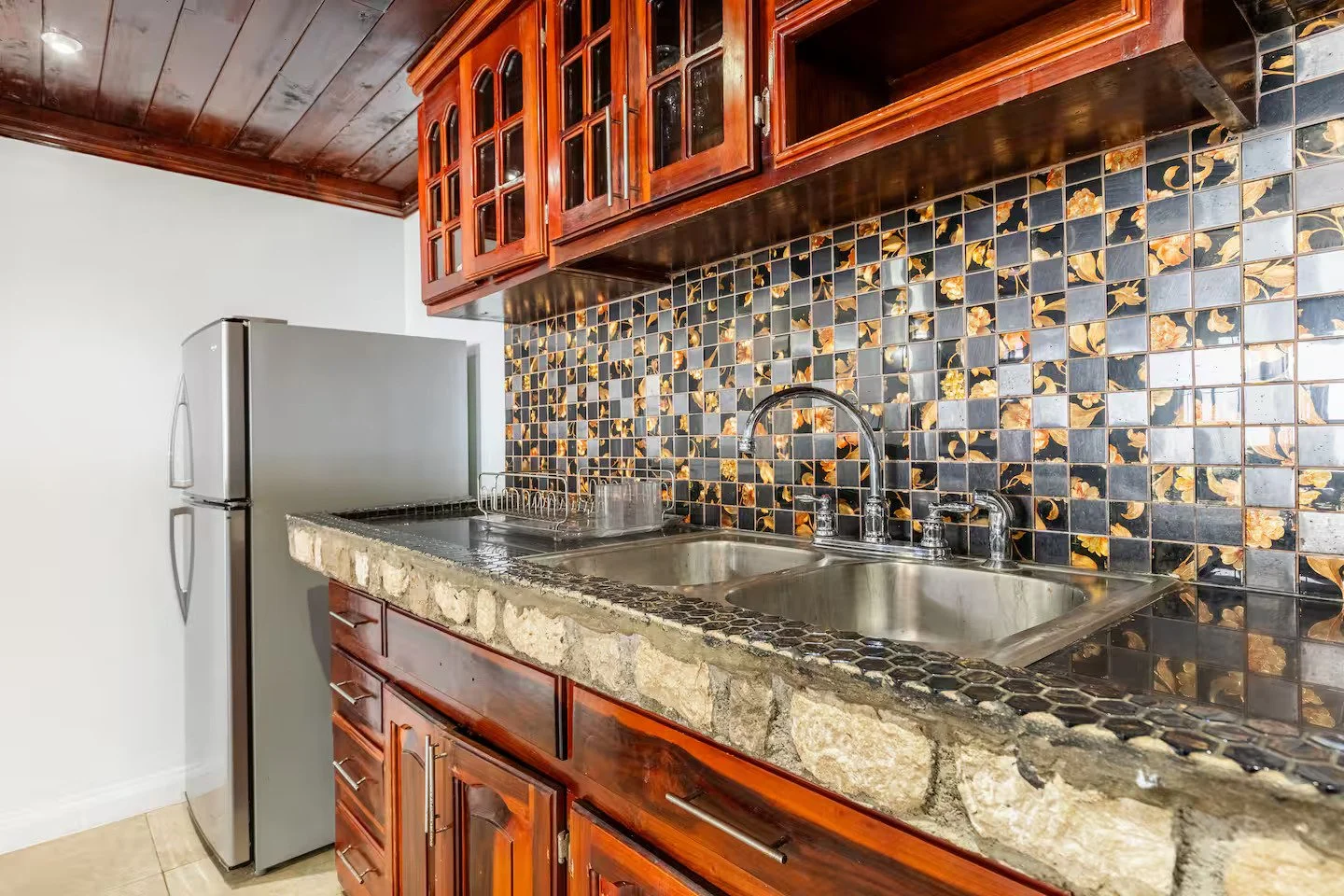 Kitchen with a double sink, floral black and gold backsplash tiles, wooden cabinets, a refrigerator, and a stone countertop.
