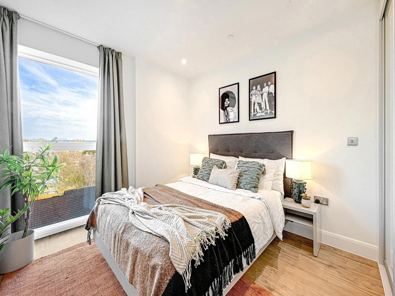 A modern bedroom with a large window, gray curtains, a bed with white bedding, multiple pillows, a black headboard, two side tables with lamps, a potted plant, and black and white framed photographs on the wall.
