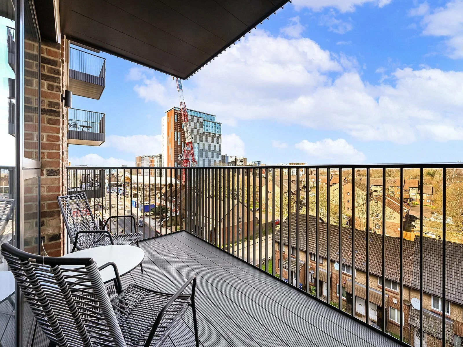 View from a balcony showing outdoor seating with a round white table and black metal chairs, overlooking a cityscape with high-rise building, construction crane, residential houses, and a partly cloudy sky.