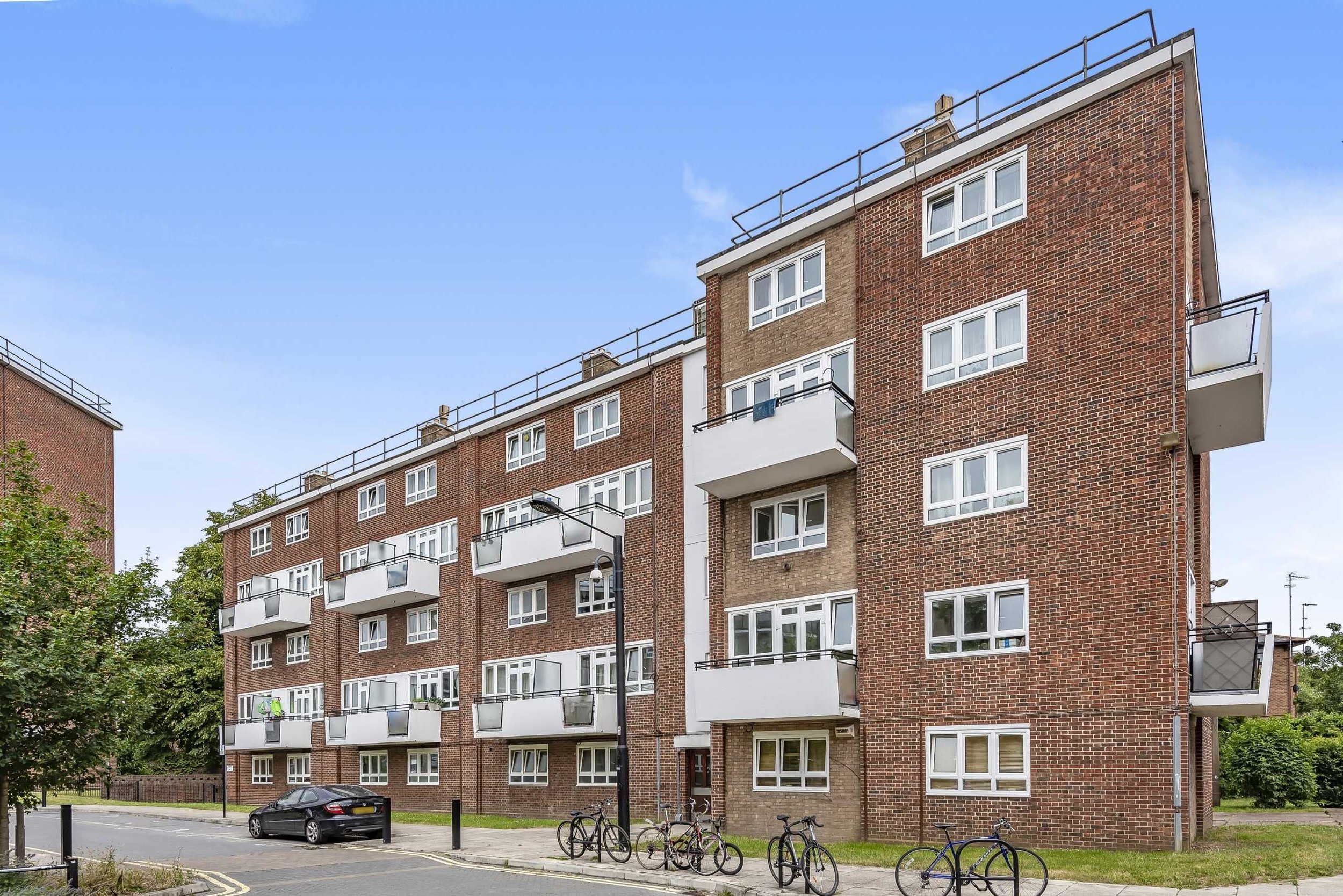 A multi-story brick apartment building with balconies, parked bicycles, a car, and a street lamp in front, under a clear blue sky.