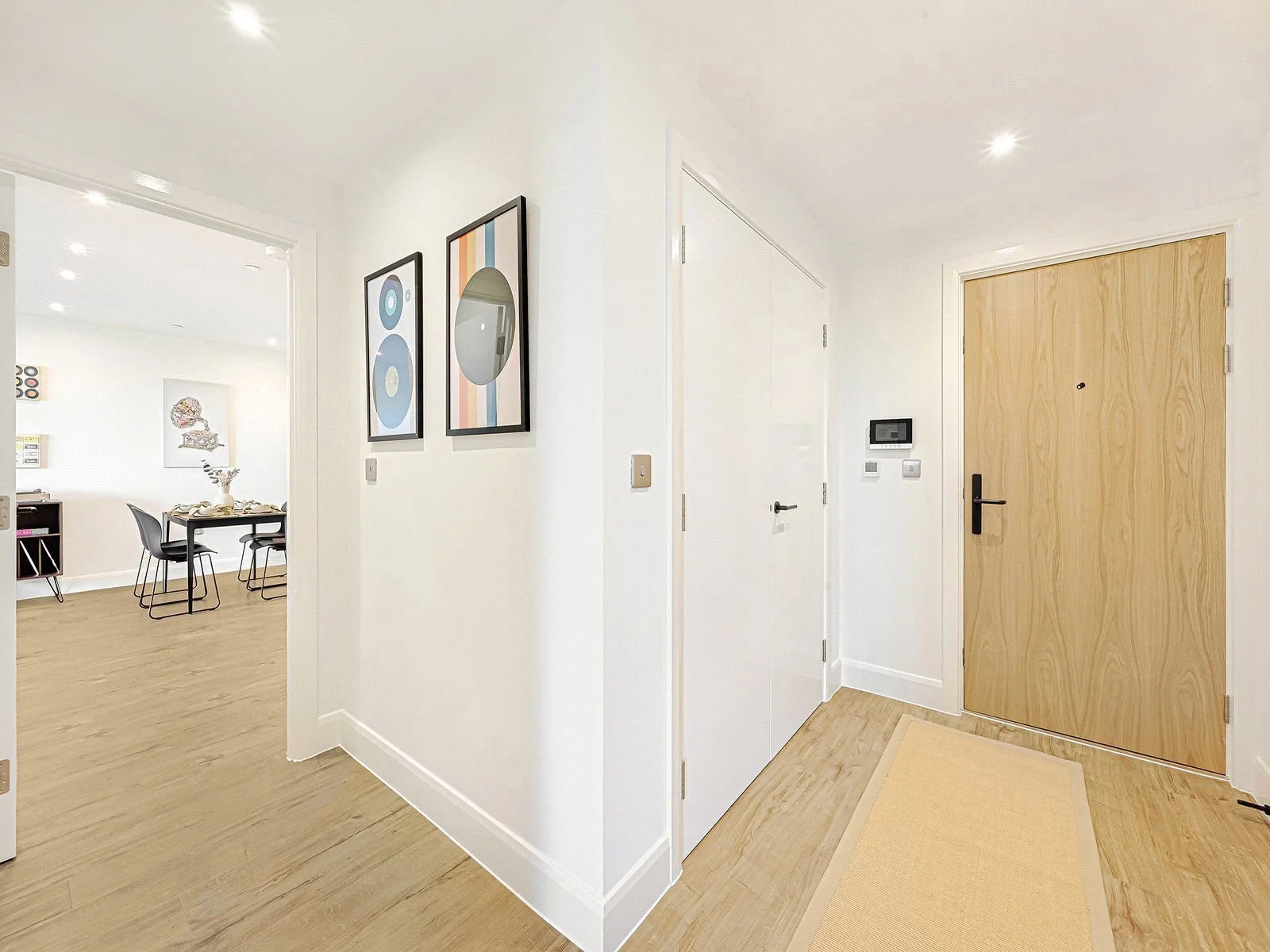 A modern apartment entryway with a wooden front door, light-colored wood flooring, a beige rug, and white walls. There are two framed abstract art pieces on the wall and adjacent rooms with dining and living areas visible.