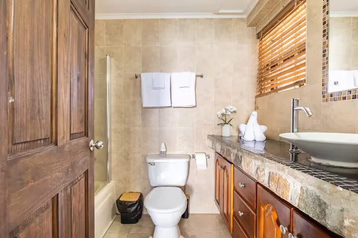 Bathroom with a wooden door, beige tiled walls, a white toilet, a trash can, a towel rack with two towels, a countertop with a sink, a flower vase, and a window with wooden blinds.