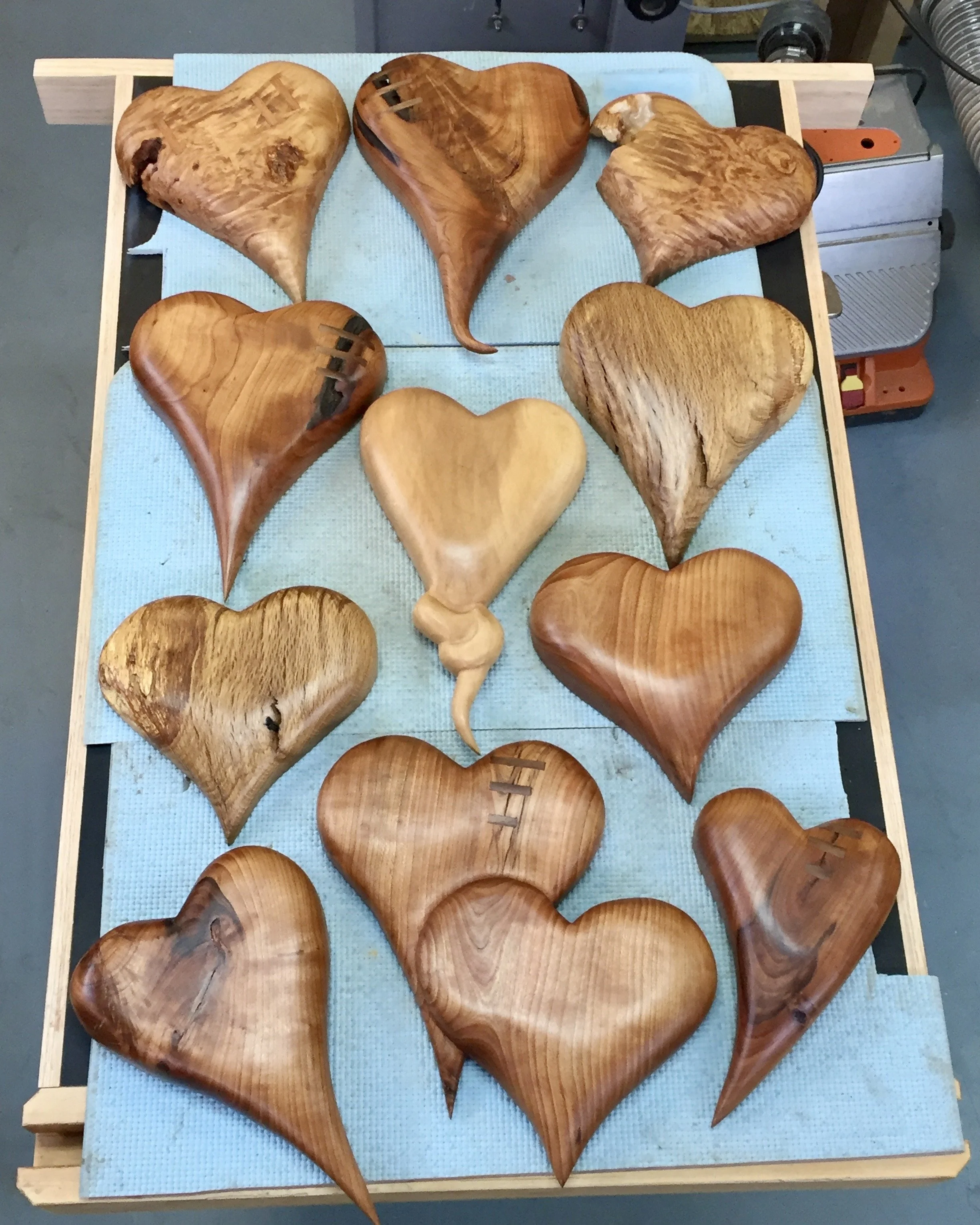 A collection of twelve wooden hearts and one wooden swirl, arranged on a blue cloth on a table in a woodworking workshop.