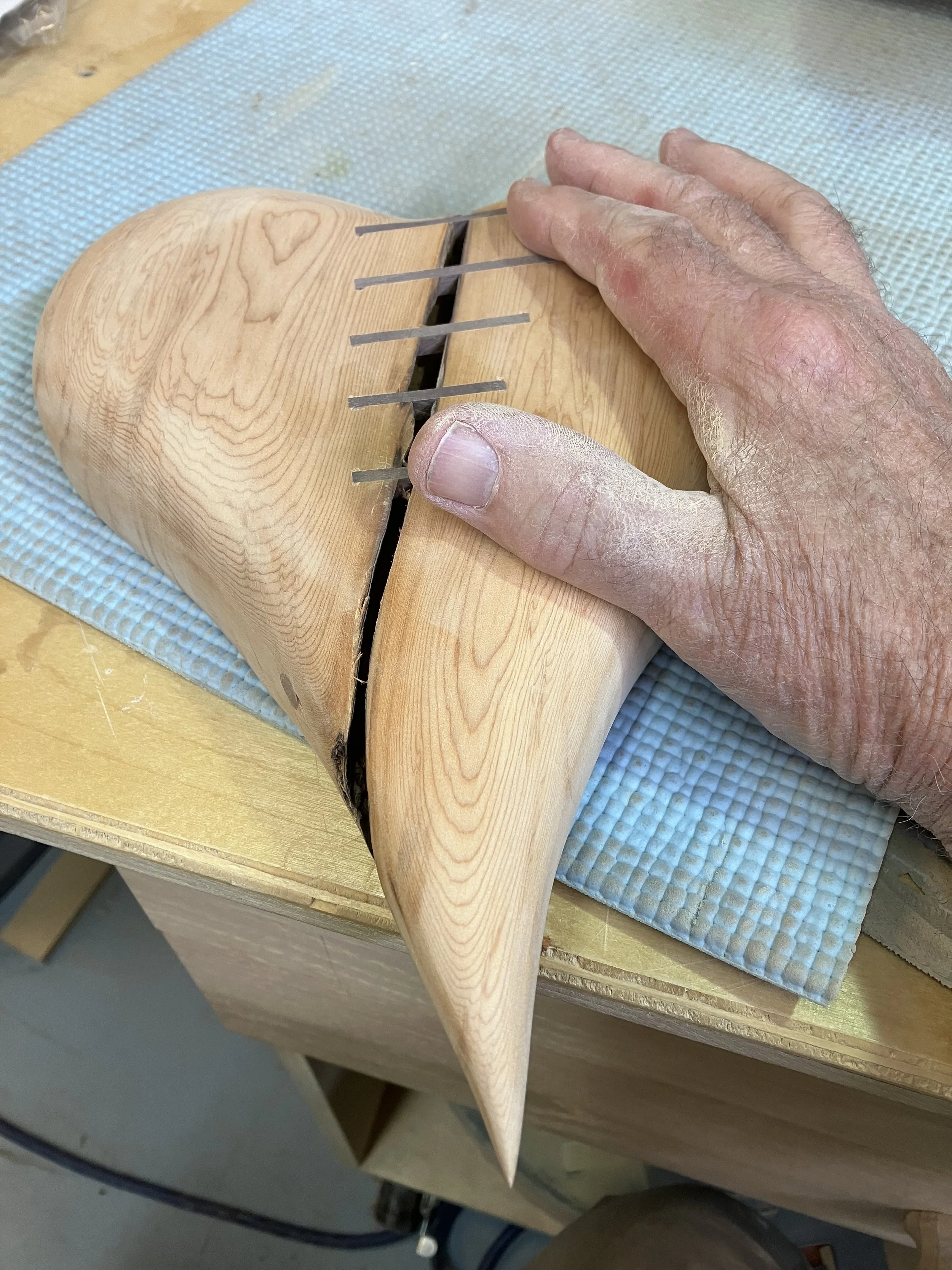 A hand with textured skin and dry patches on the fingers is holding a carved wooden object shaped like a fish jaw with metal prongs inserted into the top. The object is resting on a blue textured mat on a workbench.