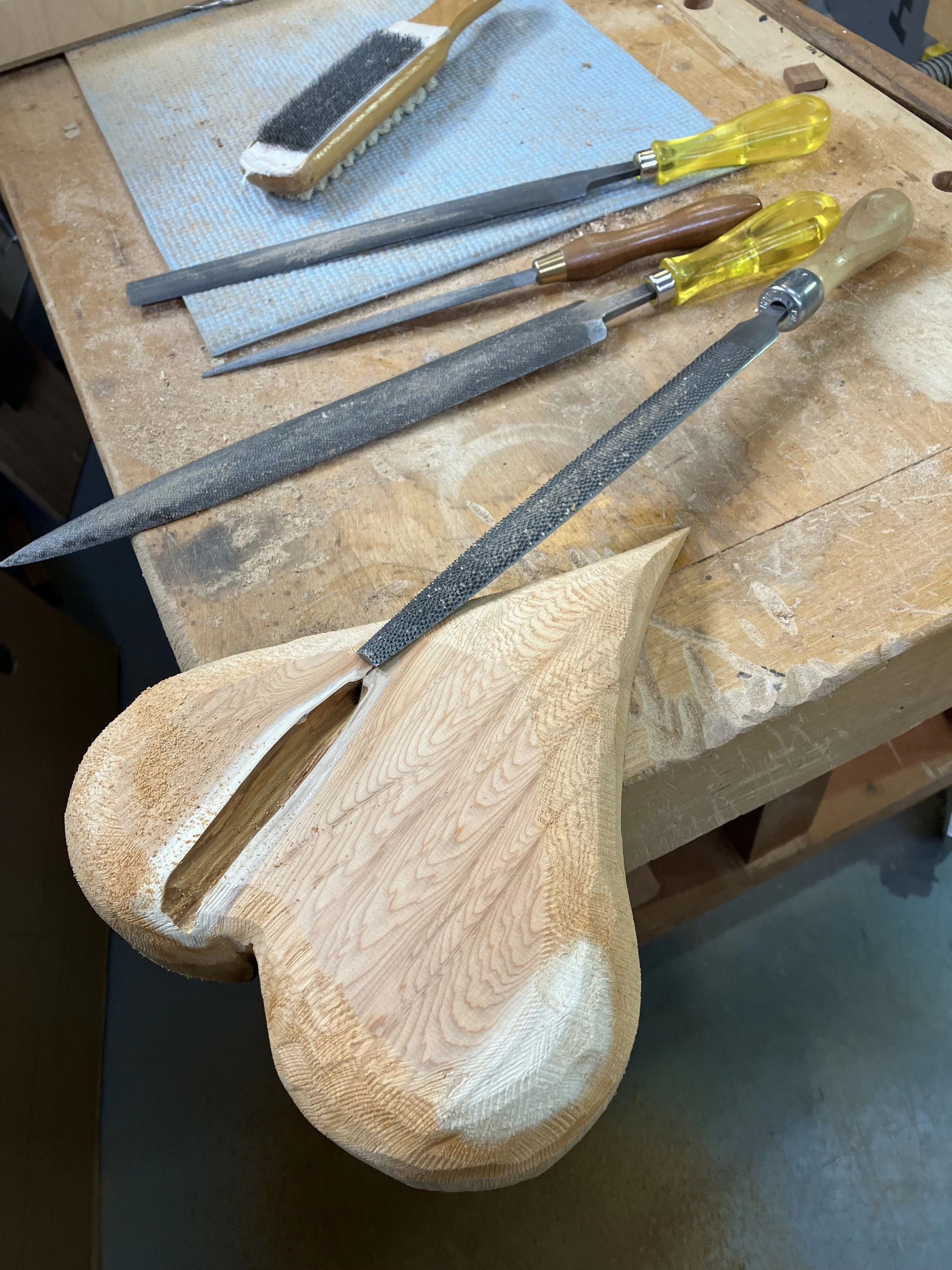 A woodworking workspace featuring a carved wooden guitar body with various files and tools on a workbench.