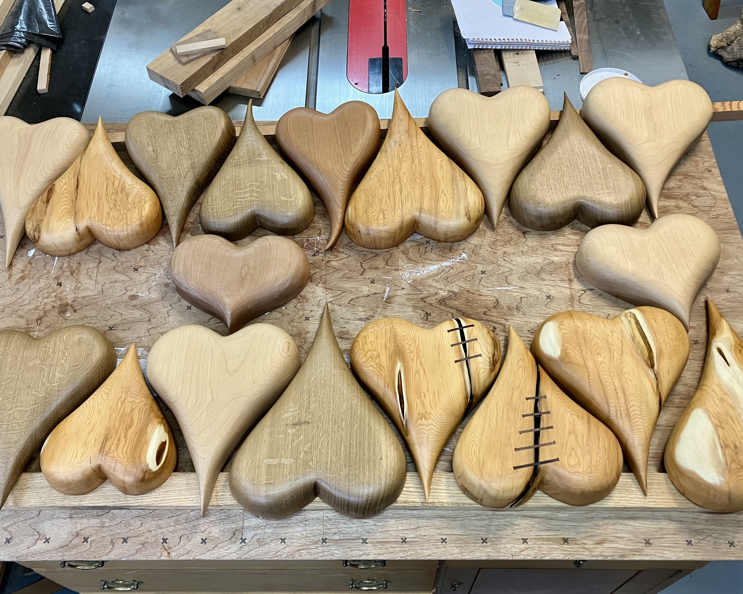 Multiple wooden heart-shaped and teardrop-shaped carvings laid out on a workbench in a woodworking shop, some decorated with carved stitches, with woodworking tools and supplies in the background.