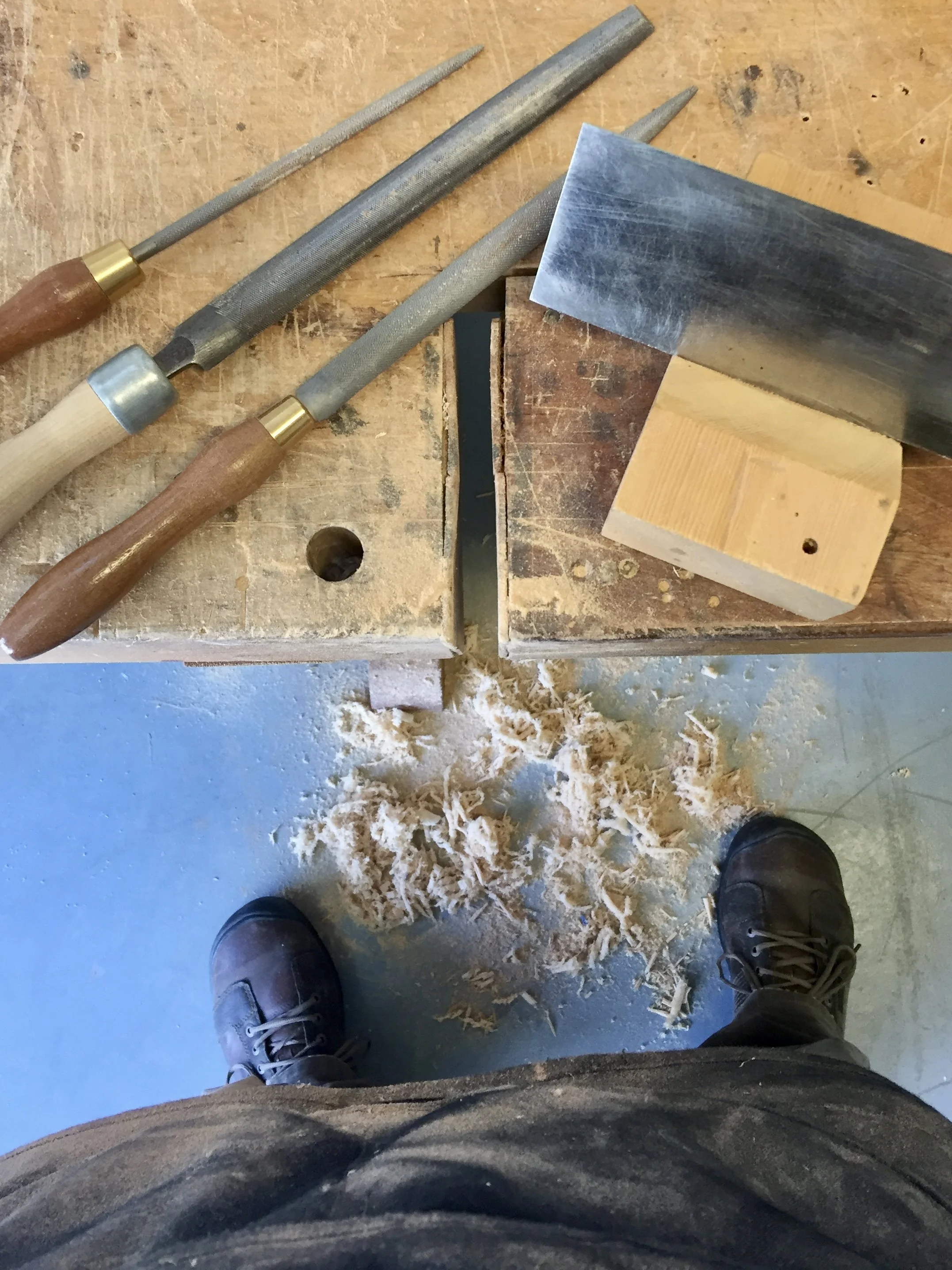 View of woodworking tools on a workbench, with wood shavings on the floor and a person wearing brown shoes standing below.