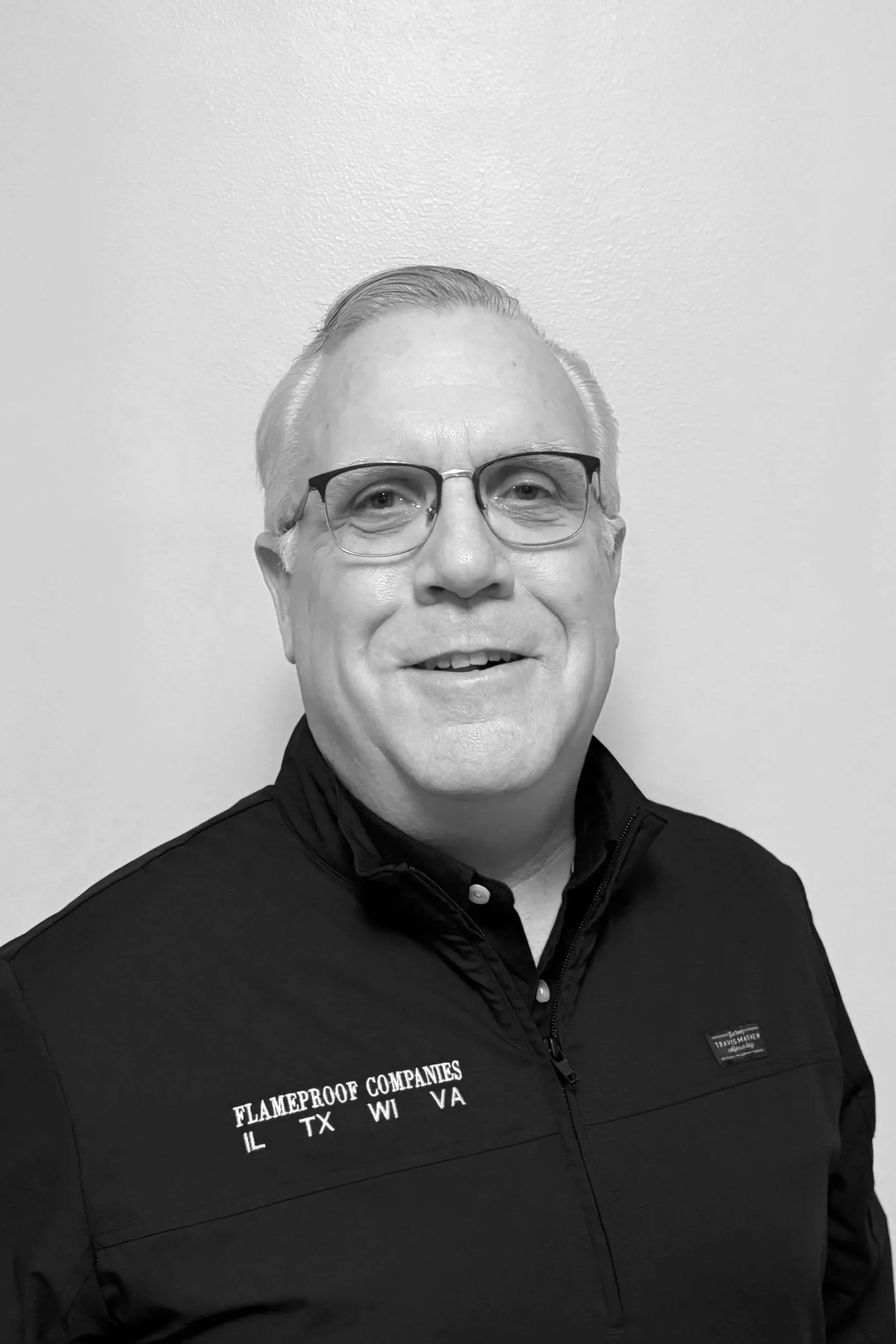 A black-and-white photo of a middle-aged man with glasses, short light-colored hair, smiling, wearing a black jacket with white text reading 'Flameproof Companies IL TX WI VA' and a high collar, standing against a plain light-colored wall.