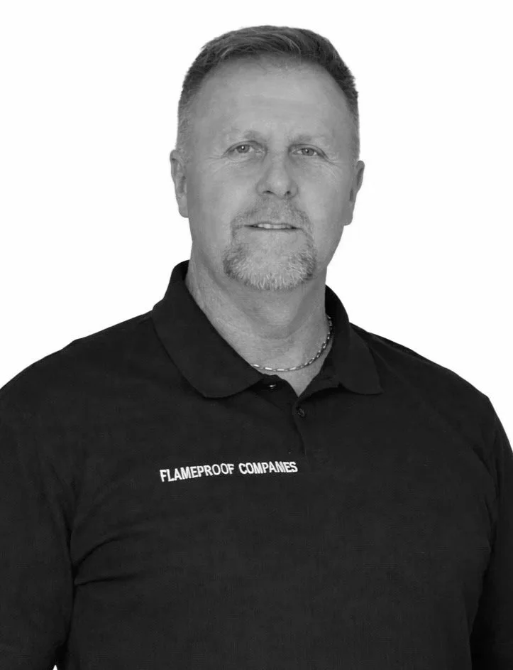 A man with short hair and a goatee wearing a black polo shirt with 'FLAMEPROOF COMPANIES' embroidered on it, standing against a plain background.