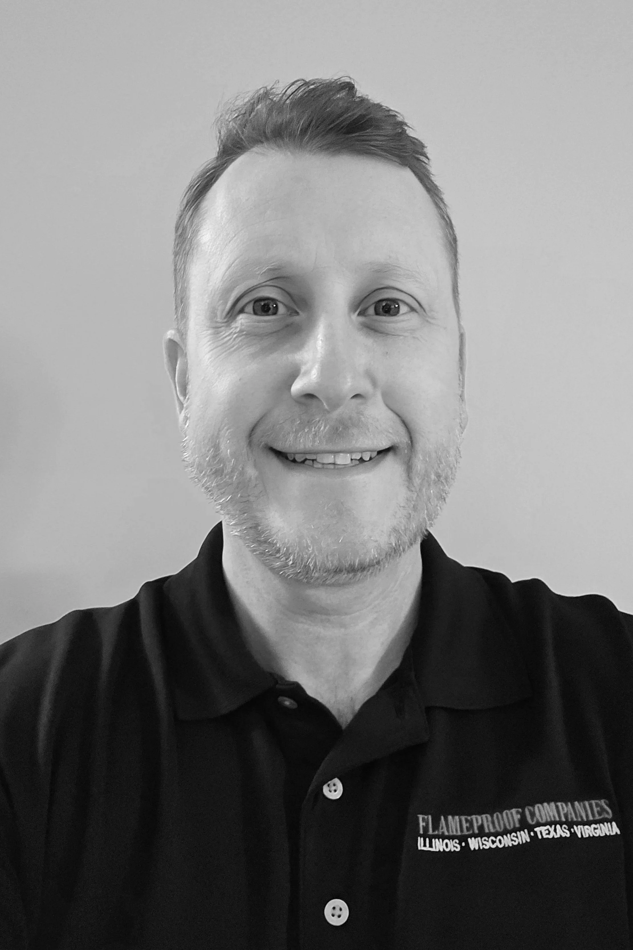 A smiling man with short hair and a beard wearing a black polo shirt that says Flameproof Companies with list of states Illinois, Wisconsin, Texas, Virginia.