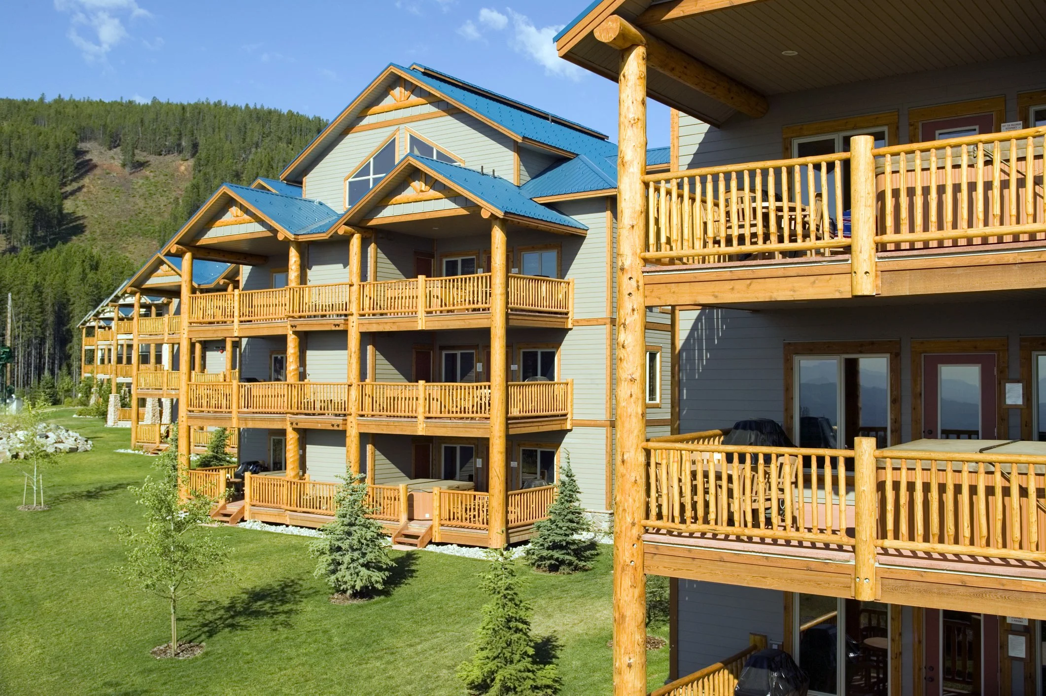 Multi-story residential building with wooden balconies and blue metal roofs, surrounded by green grass, small trees, and mountainous landscape in the background.
