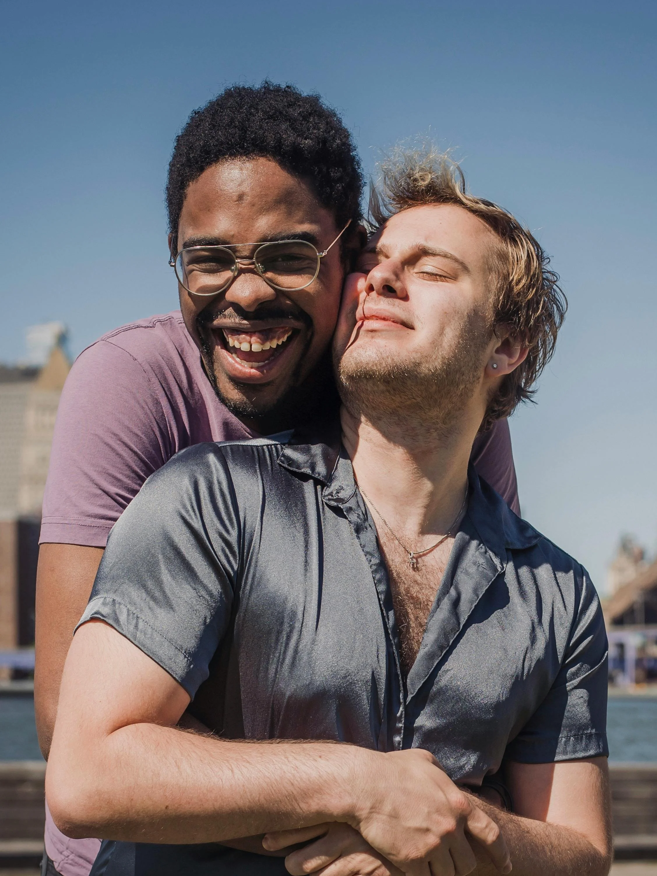 Two men hugging outdoors against a clear blue sky, smiling and enjoying each other's company.
