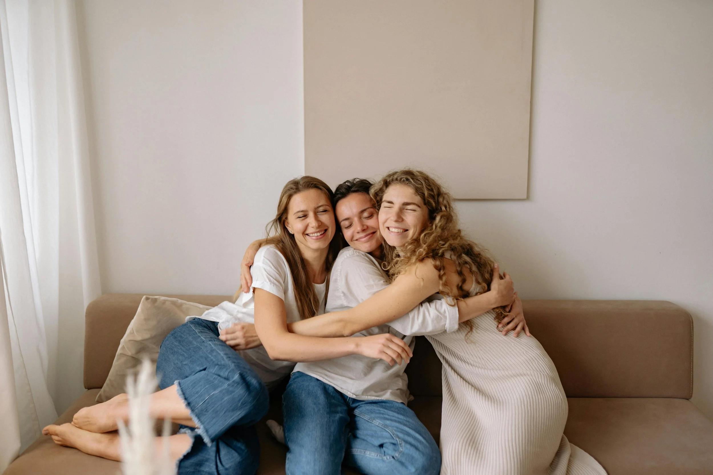 Three women hugging and smiling on a beige couch in a well-lit room after Dallas EMDR session.