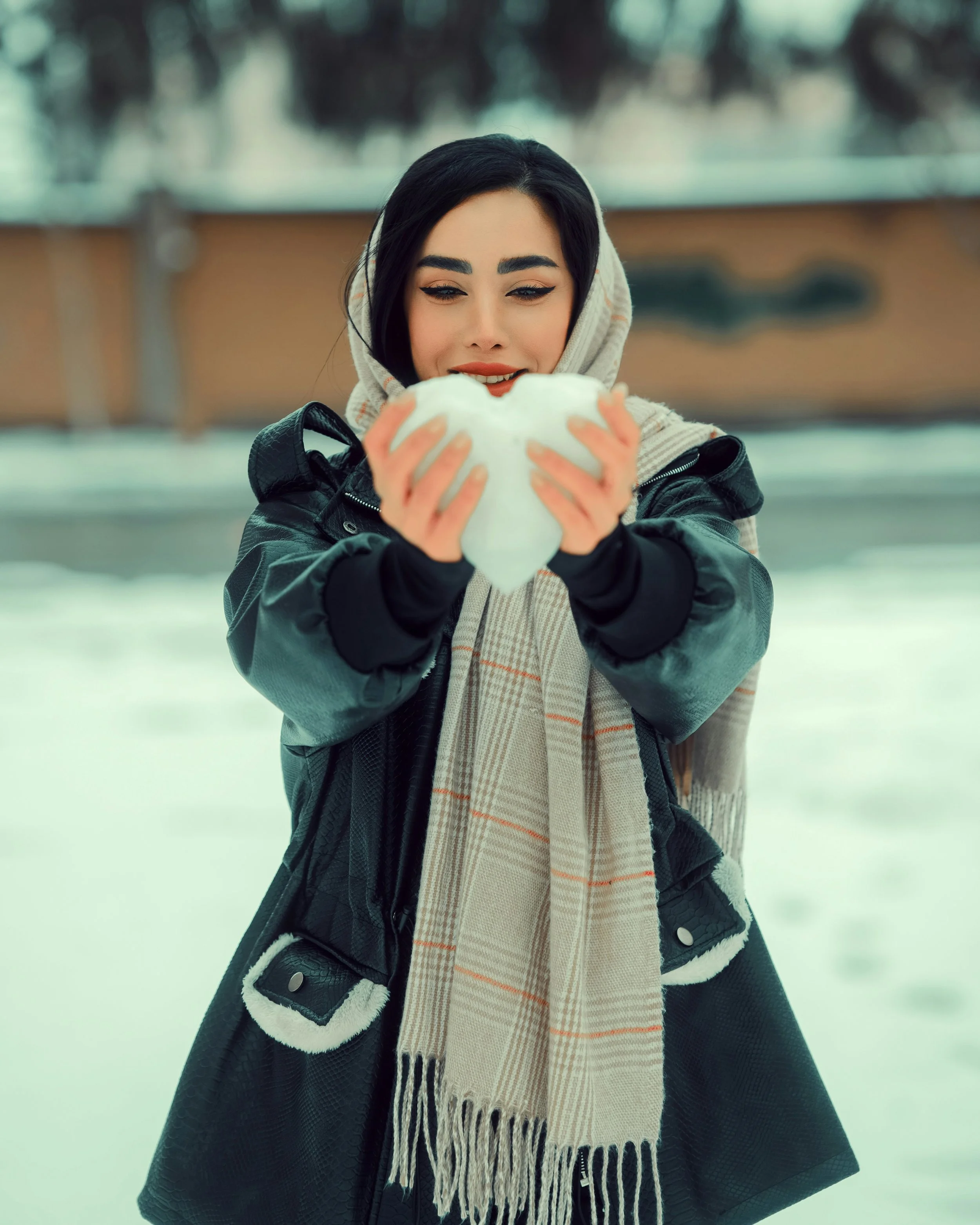 An adult survivor of childhood negelct in winter clothing holding and looking at a heart-shaped snowball outdoors in a snowy setting.