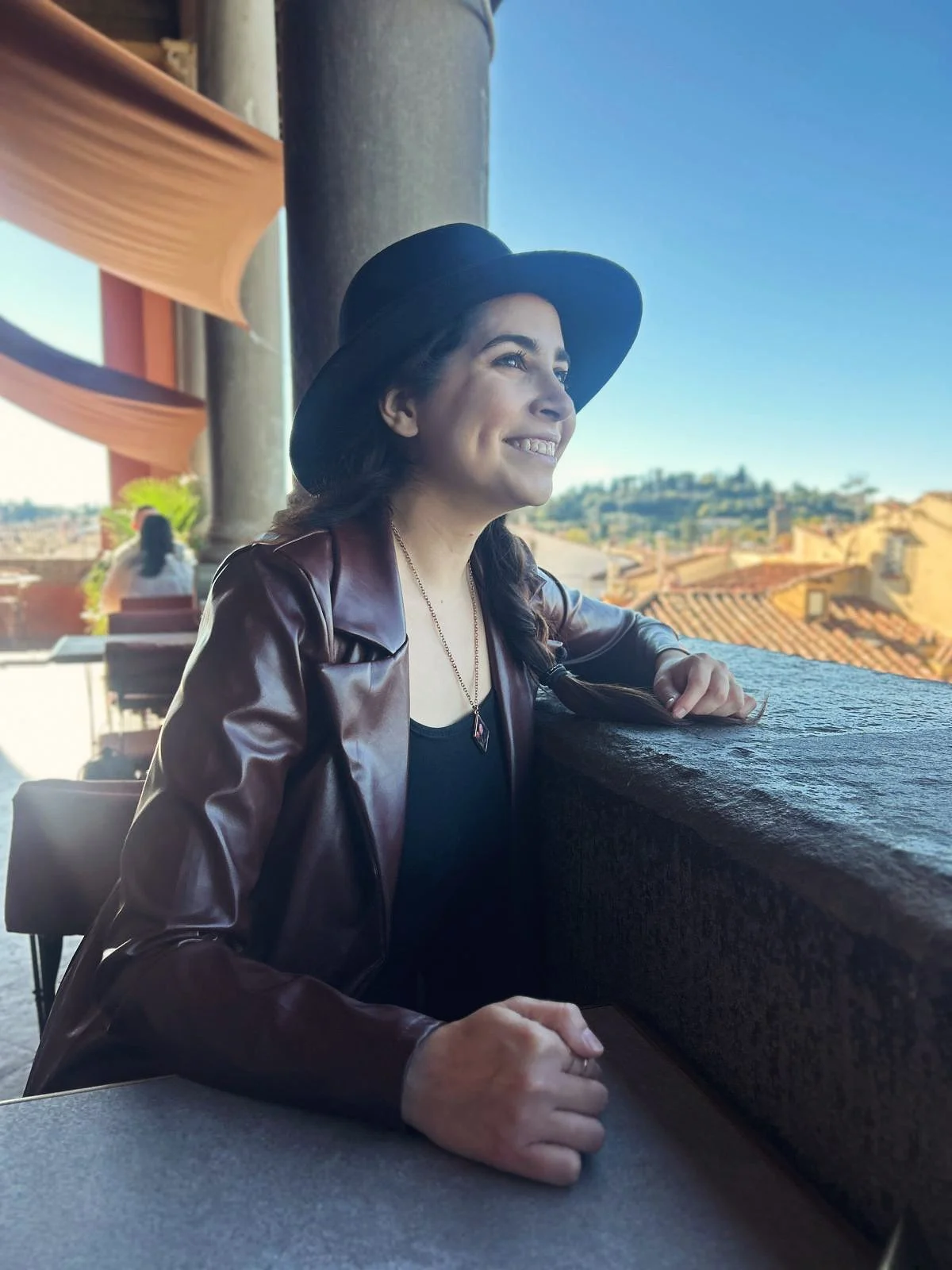 A Dallas therapist sitting outdoors at a table, wearing a black hat, a leather jacket, and a necklace. She is smiling and looking into the distance with a scenic view of rooftops and a hill in the background.