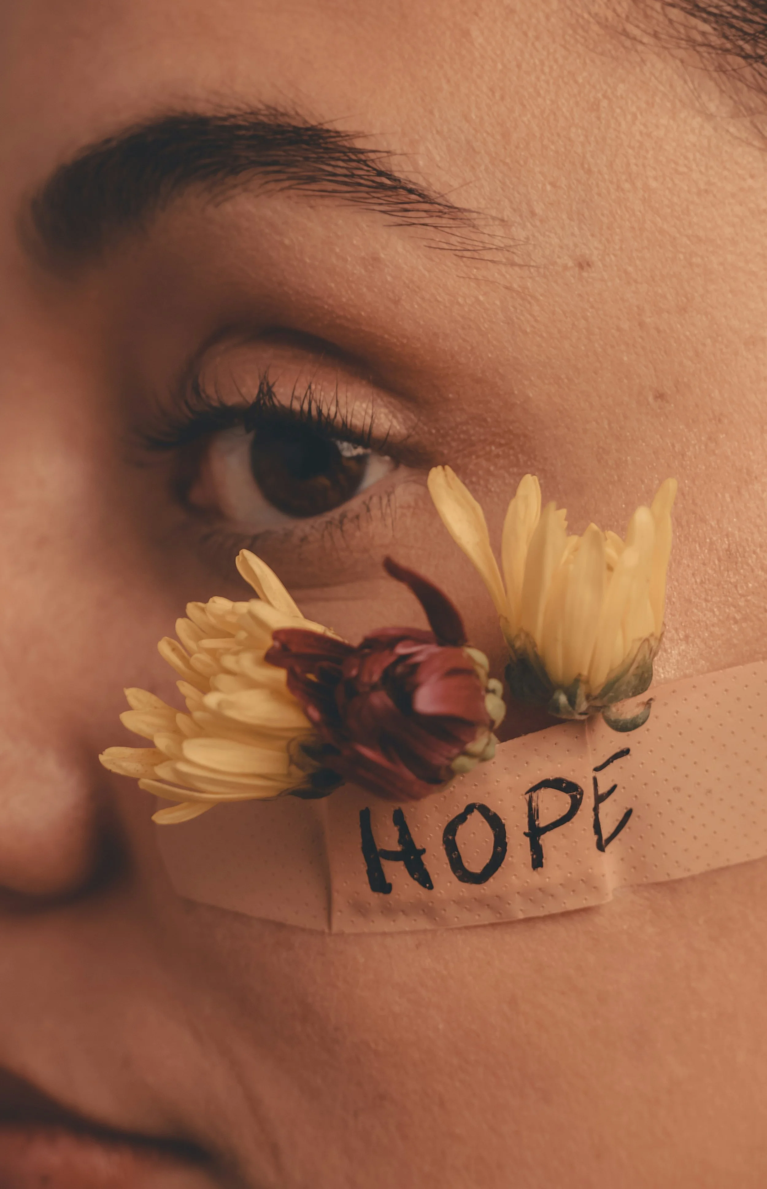 Close-up of a female trauma survivor's face with a bandage on their cheek labeled 'HOPE', decorated with yellow and pink flowers.