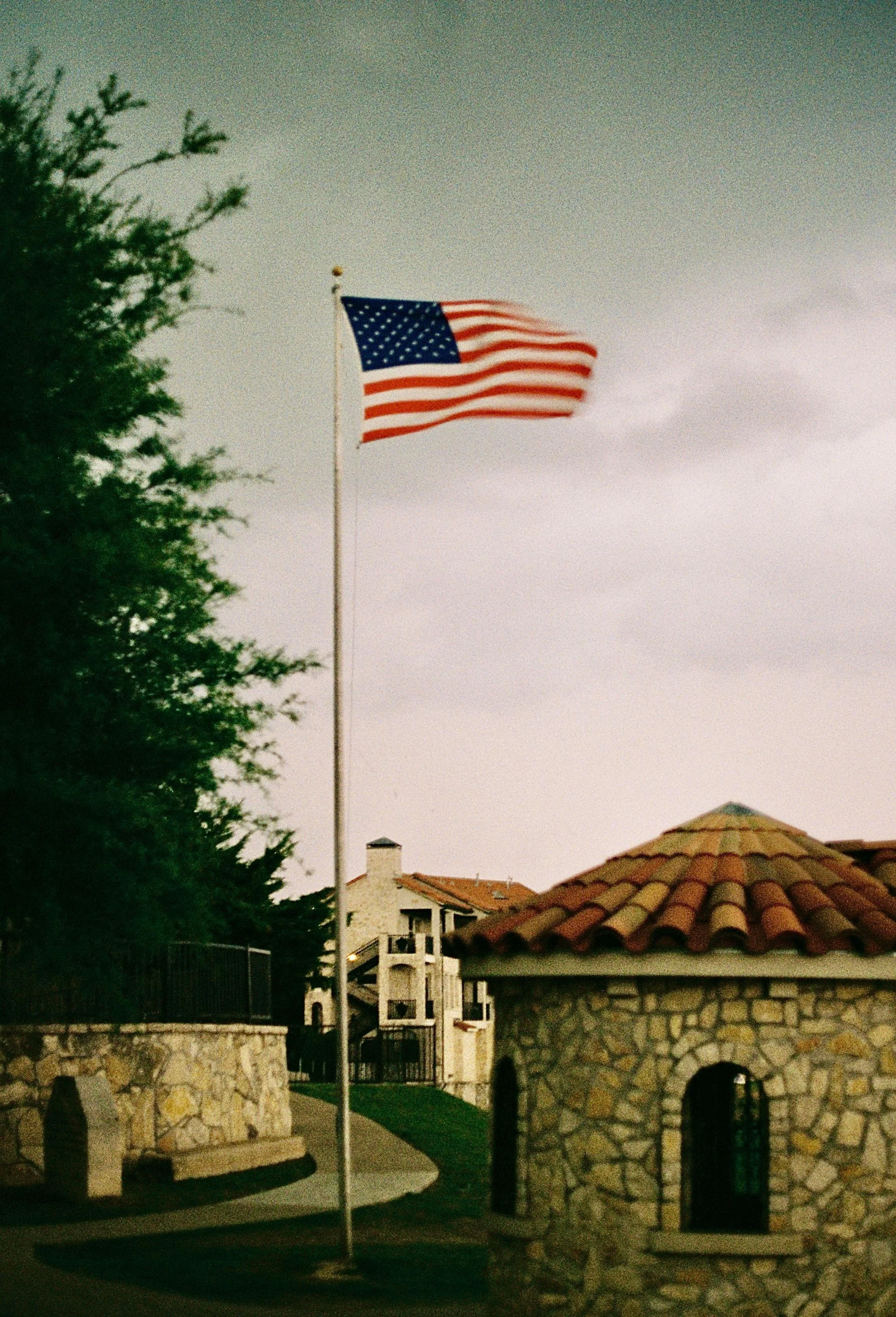 American flag flying on a flagpole in front of a stone building in Texas with a tiled roof, with trees and a residential building in the background.