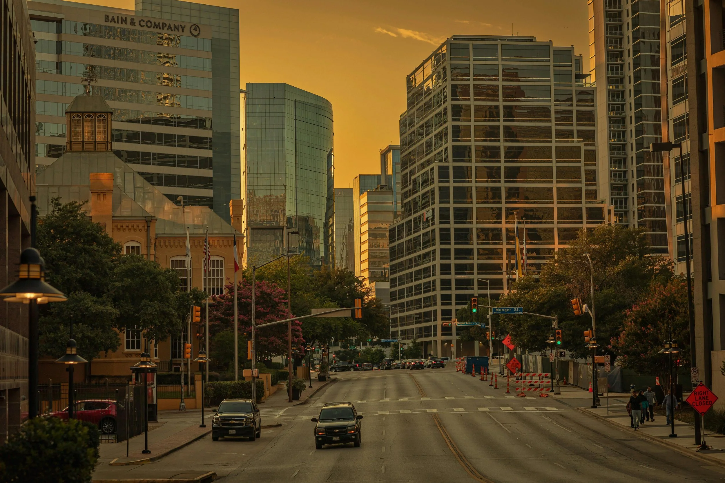 Uptown Dallas street at sunset with tall glass buildings, trees, cars, and pedestrians.