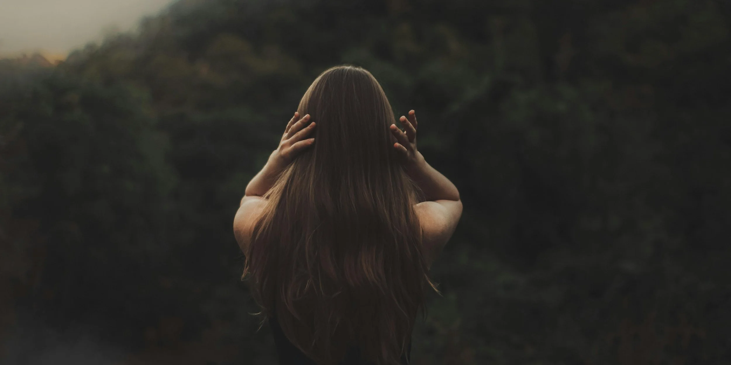 A female childhood trauma survivor with long brown hair, viewed from behind, holding her head with both hands in a natural outdoor setting during sunset or dusk.