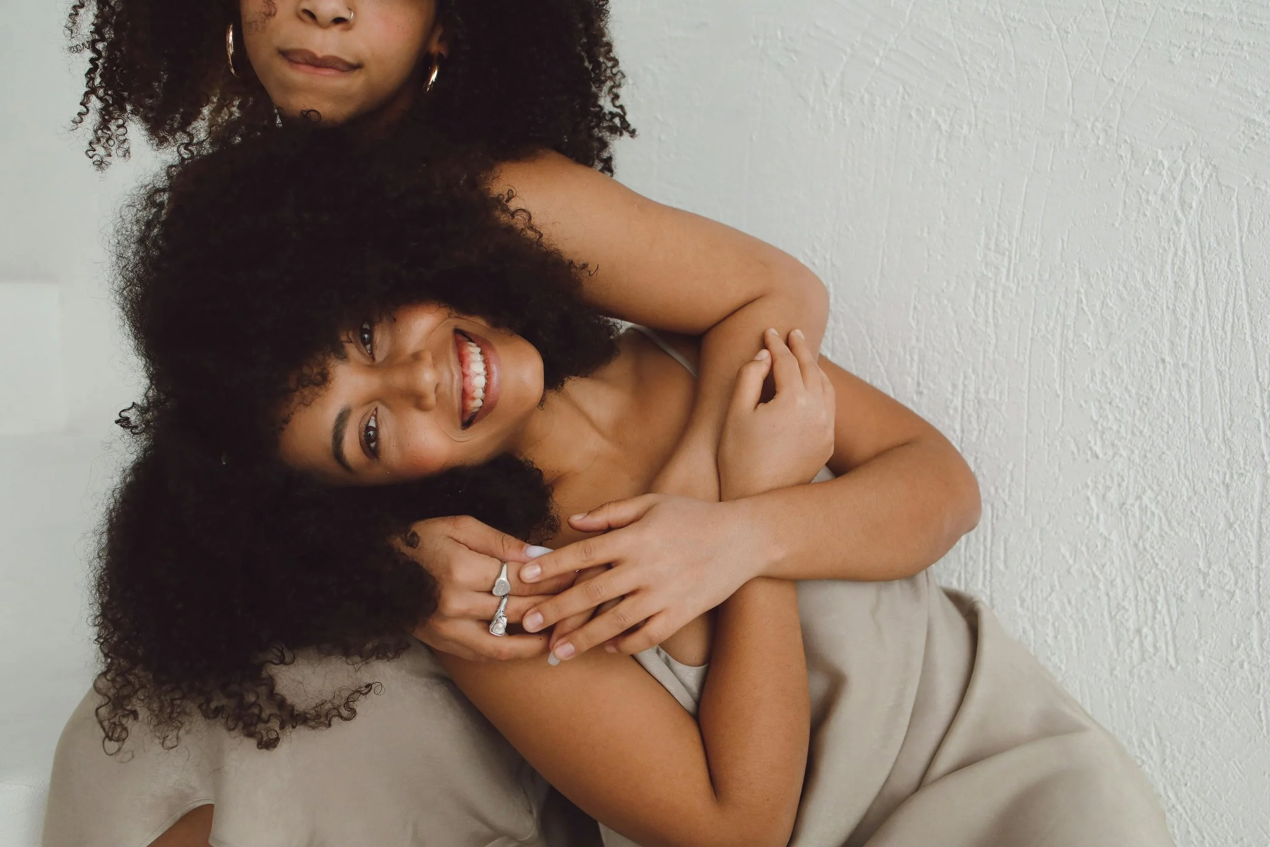 Two female trauma survivors with curly hair smiling and embracing each other against a white textured wall.
