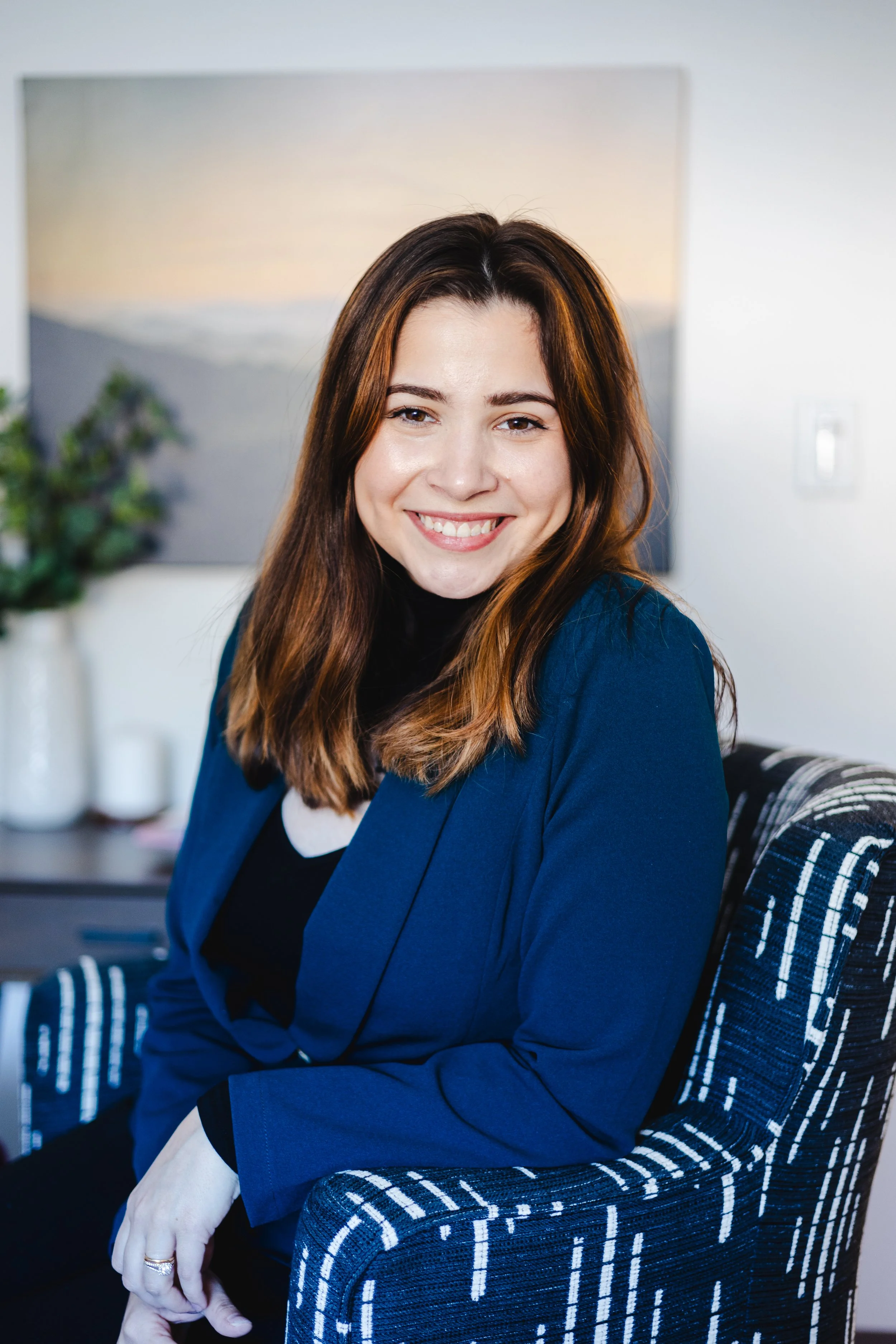 A Texas mental health counselor with brown hair smiling, sitting on a patterned chair in a modern North Dallas therapy office with a painting and a plant in the background.