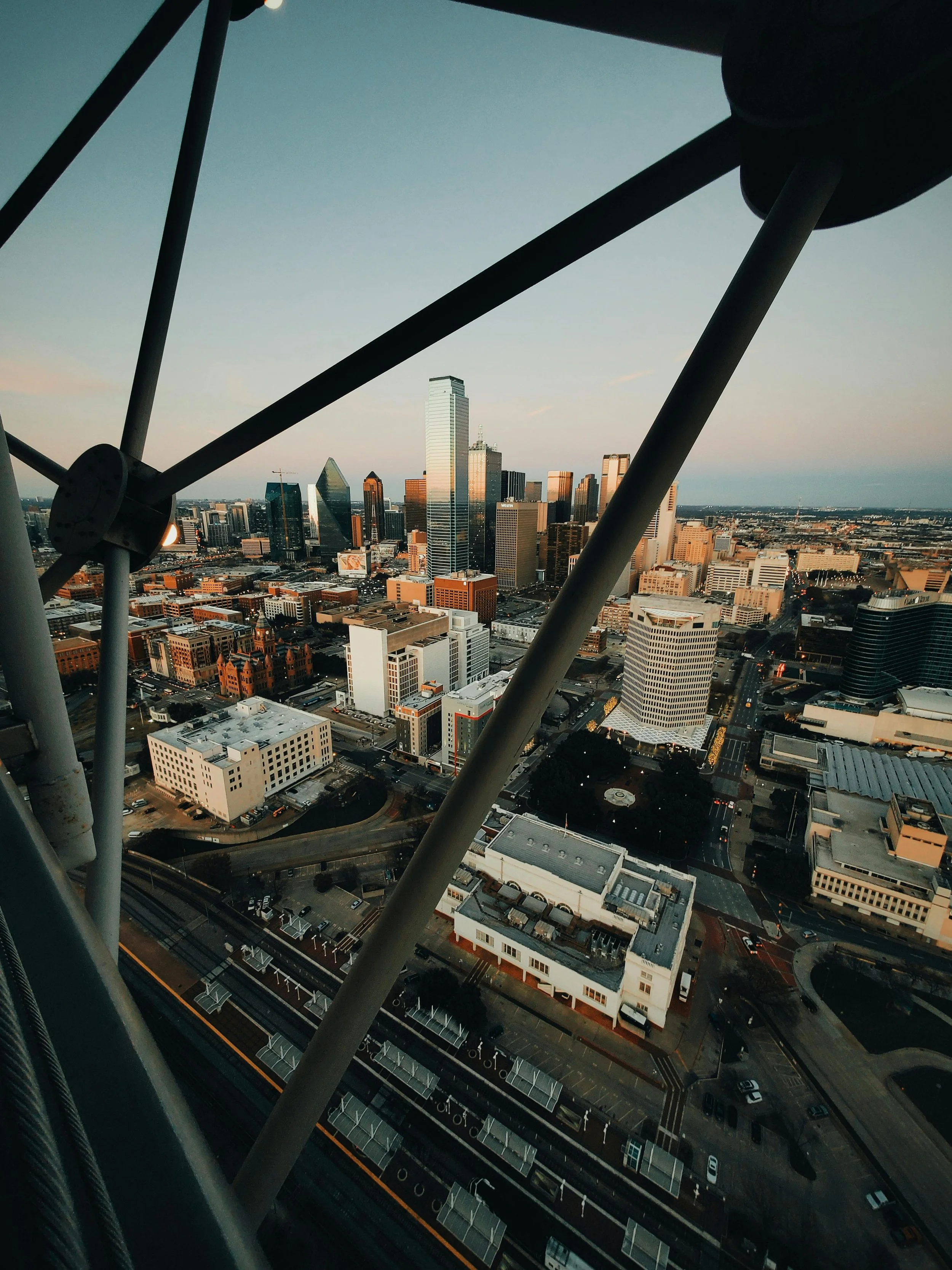 Cityscape of Uptown Dallas, Texas seen from a high vantage point at sunset, with a construction crane in the foreground and tall skyscrapers in the background.