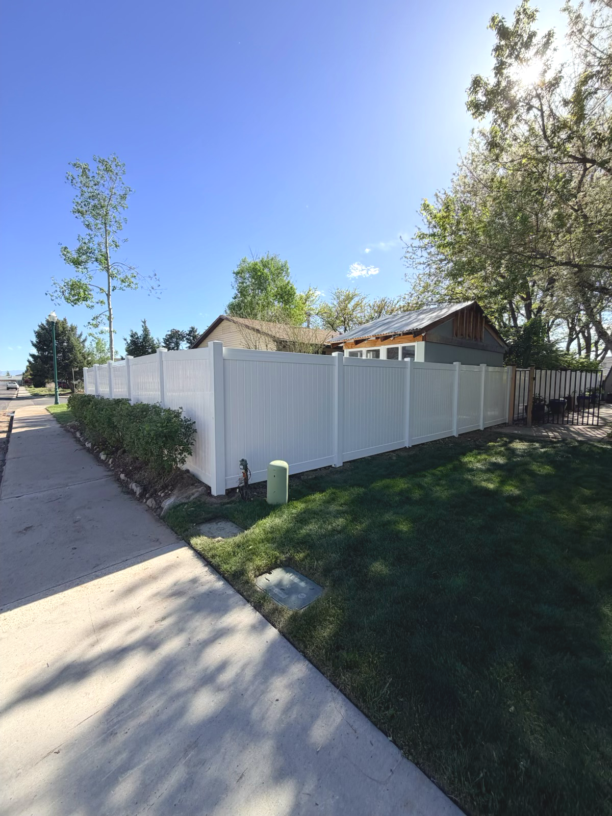 A residential neighborhood scene with a new white vinyl privacy fence surrounding a backyard. The fence runs along a sidewalk and over a grass lawn, with trees and a house in the background under a clear blue sky.