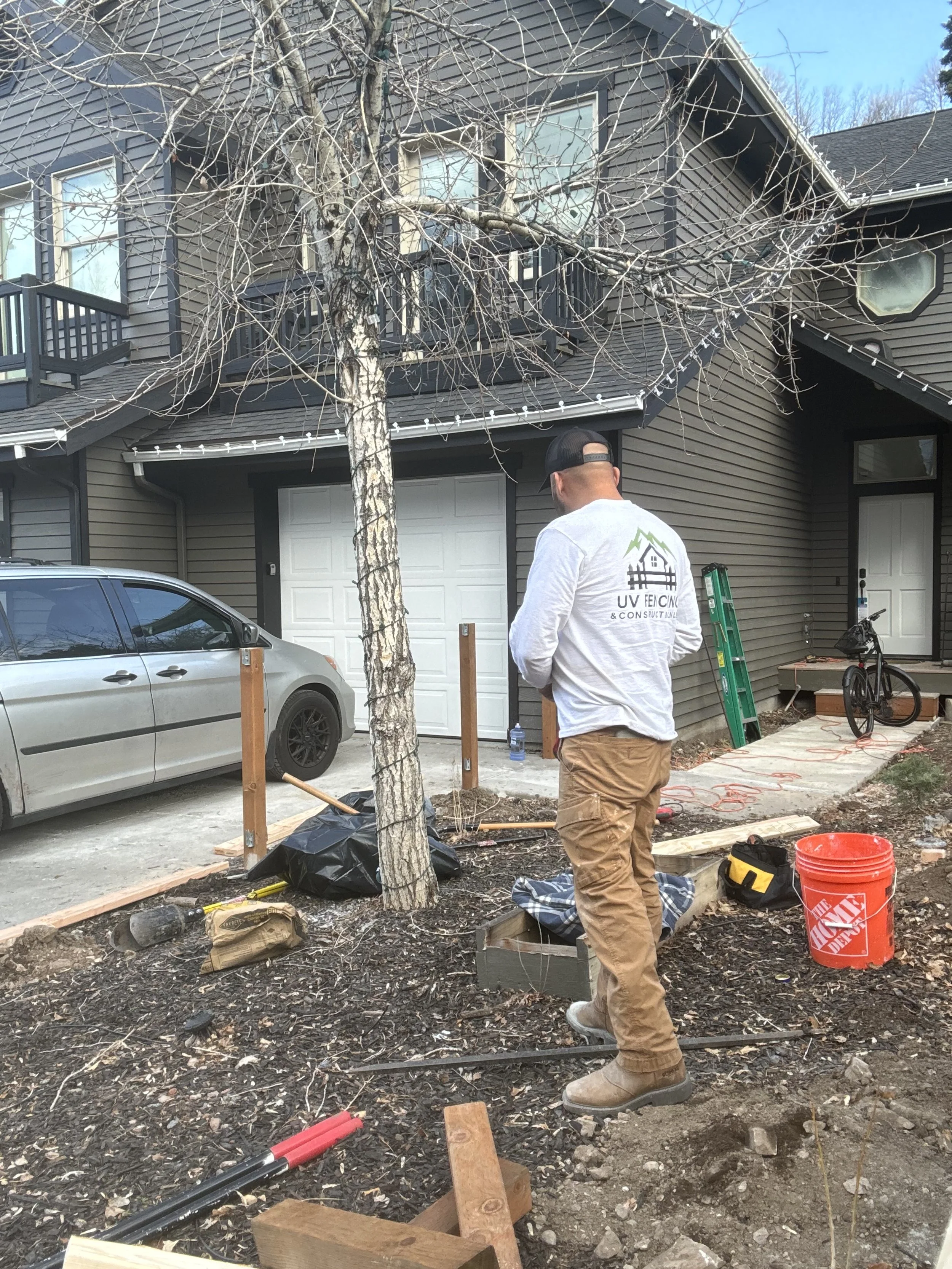 A person standing in a backyard garden area with a house in the background, working on landscaping or construction, with tools and equipment around.