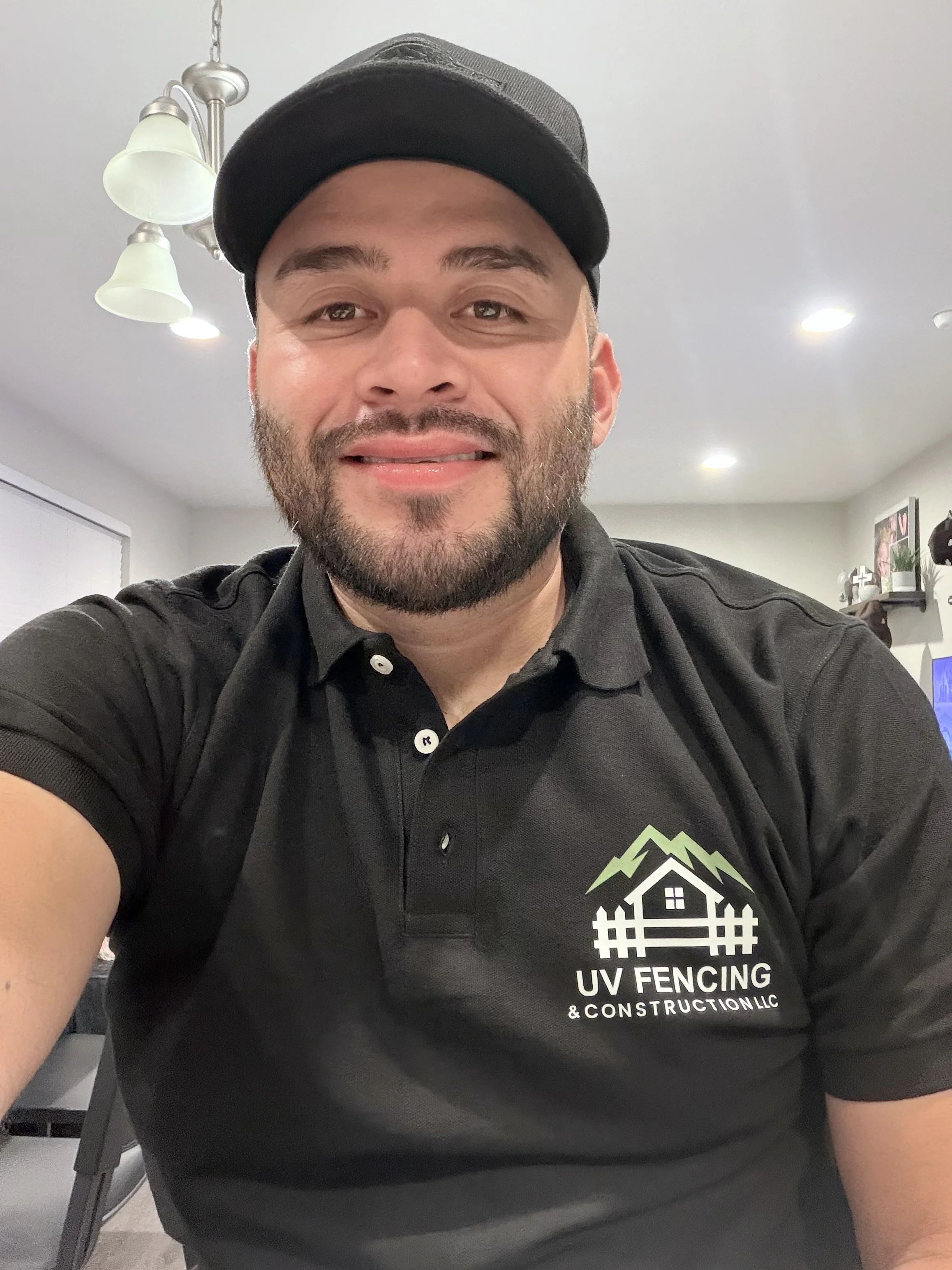 A man wearing a black UV Fencing & Construction LLC polo shirt and a black cap, smiling inside a house with ceiling lights and wall decorations.