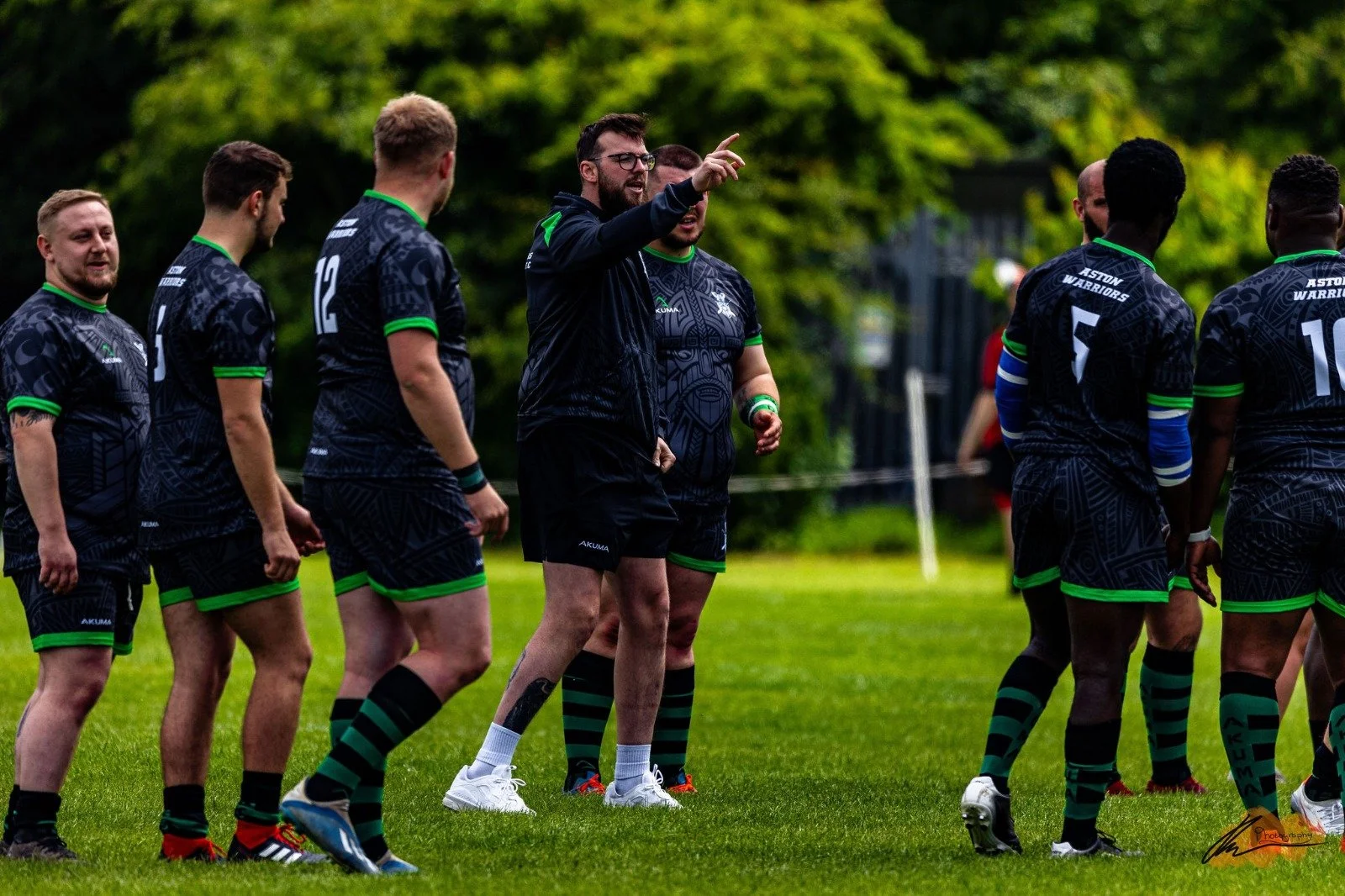 A group of rugby players in black jerseys with green accents standing on a grassy field, with one coach or team leader talking and gesturing towards them.
