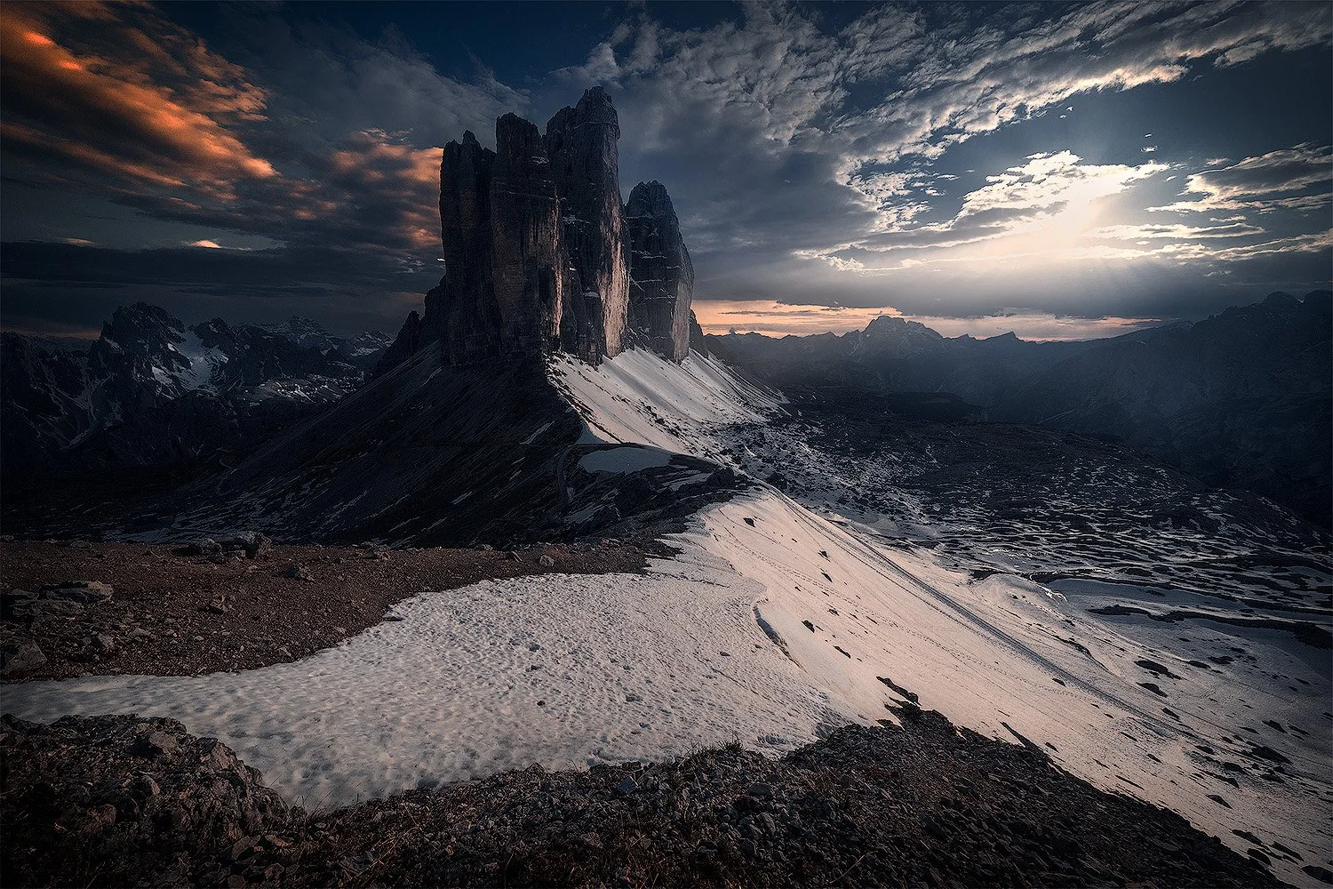 Berglandschaft mit Felsen, schneebedecktem Gelände und dunklen Wolken bei Sonnenuntergang. Dolomiten, Tre Cime