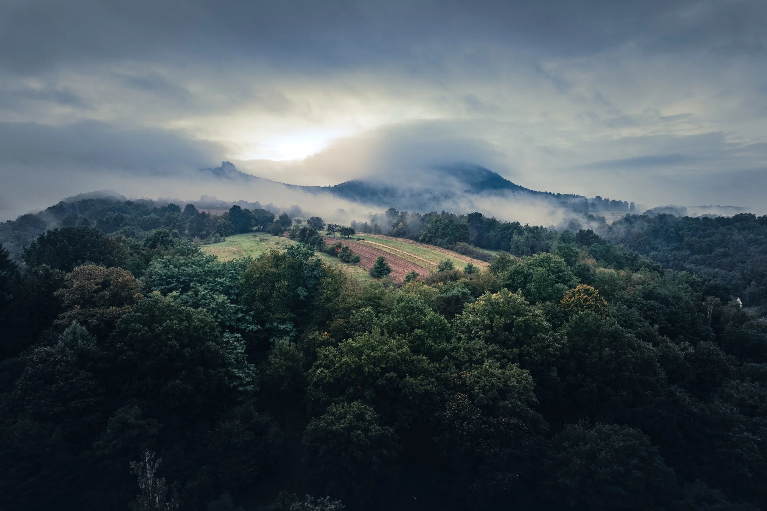 Ein nebliger Berglandschaft mit bewaldeten Hügeln und einer bewölkten Himmelsszene bei Sonnenaufgang oder Sonnenuntergang.