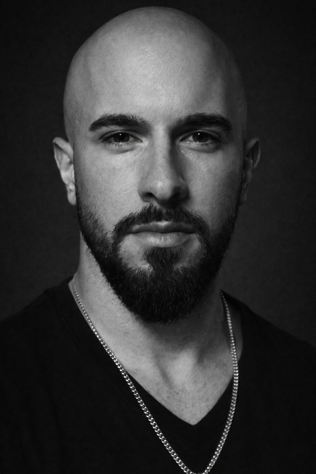 Schwarz-weiß Portrait eines Mannes mit Bart und glattem Kopf, wearing a V-neck shirt und einer Silberkette.