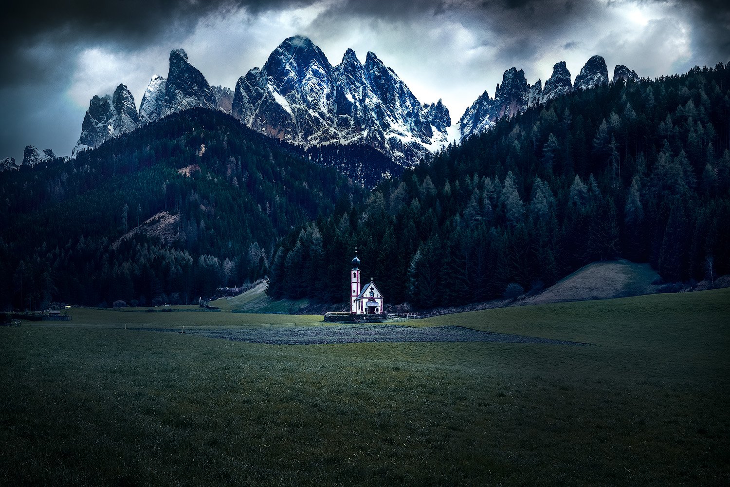 Eine kleine weiße Kirche mit einem schwarz-gelben Turm steht in einem grünen Feld vor einem dichten Nadelwald, im Hintergrund hohe schneebedeckte Berge unter bewölktem Himmel.