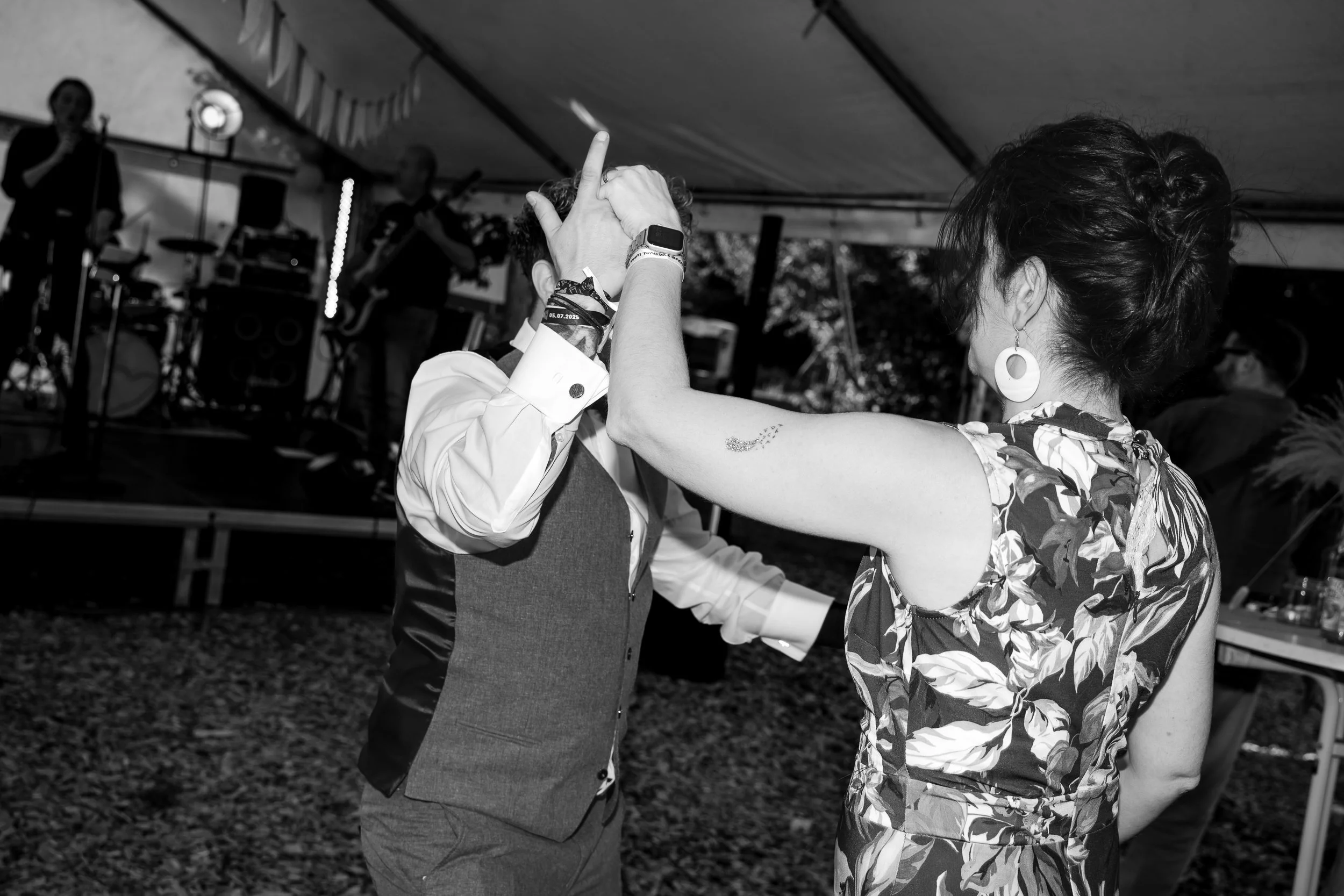 Ein Paar tanzt auf einer Party unter einem Festzelt, im Hintergrund eine live Band. Die Frau trägt ein Blumenmuster Kleid und große Ohrringe, der Mann ein Hemd und Weste.