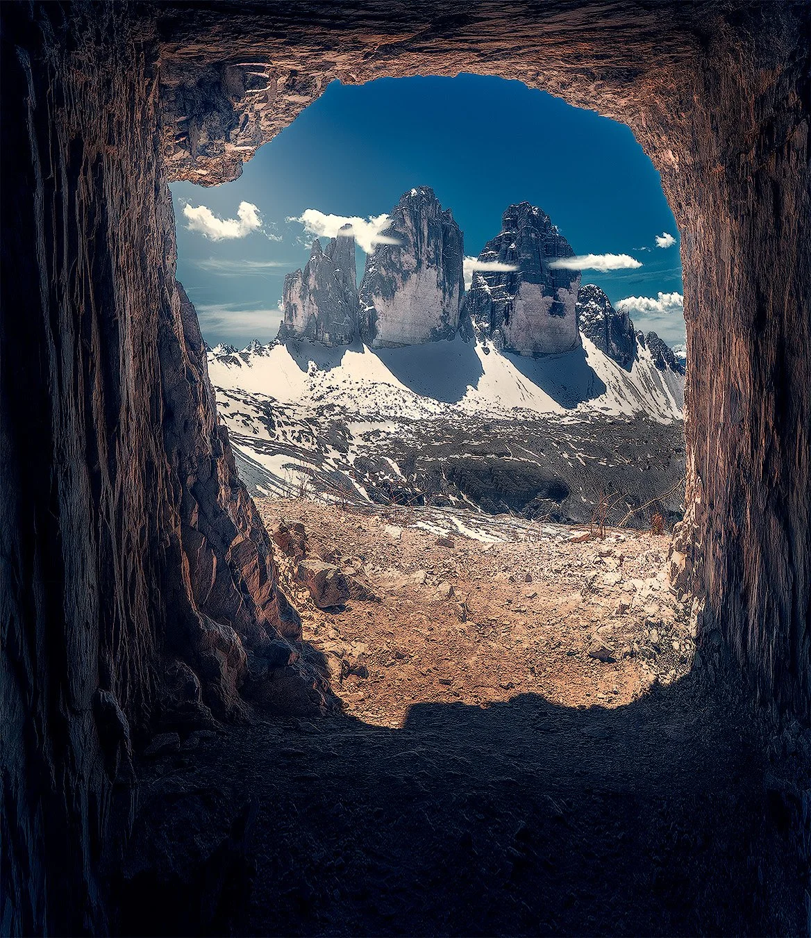 Blick aus einer Höhle auf schneebedeckte Berge und einen blauen Himmel mit Wolken. Tre Cime, Dolomiten