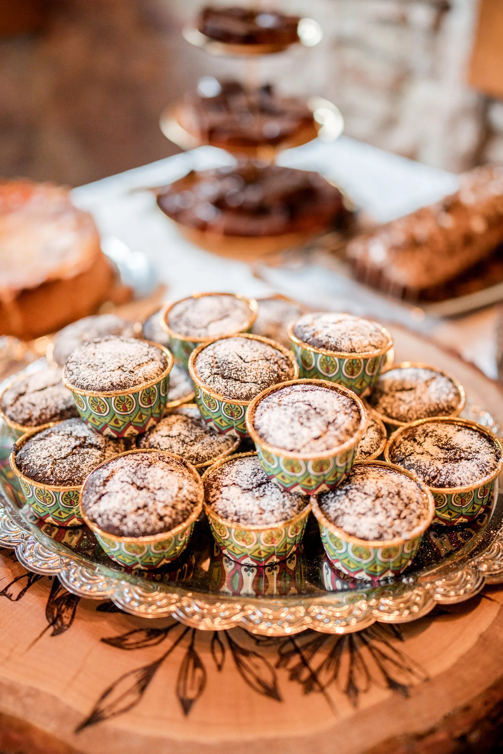Bunte Schüssel mit kleinen Muffins, die mit Puderzucker bestäubt sind, auf einem Silbertablett, im Hintergrund sind Kuchen und Torten sichtbar.