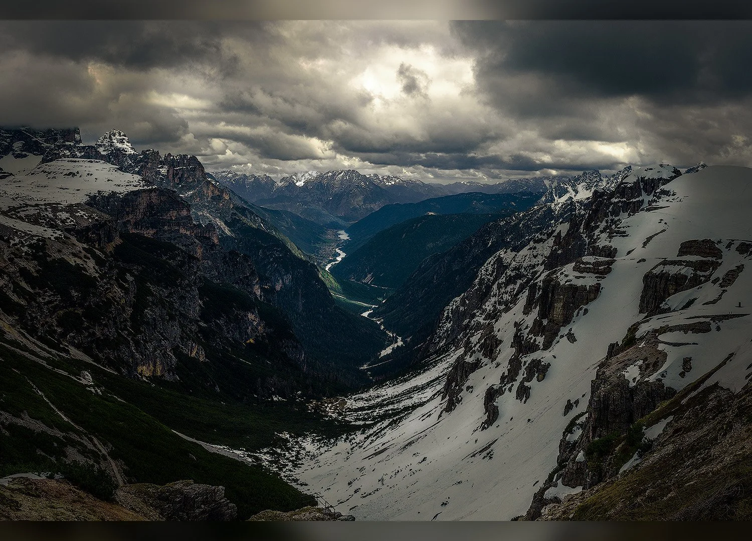 Berglandschaft mit schneebedeckten Gipfeln im Nebel und Wolken, tiefer Talfluss, grüne Bereiche und dunkle Wälder.