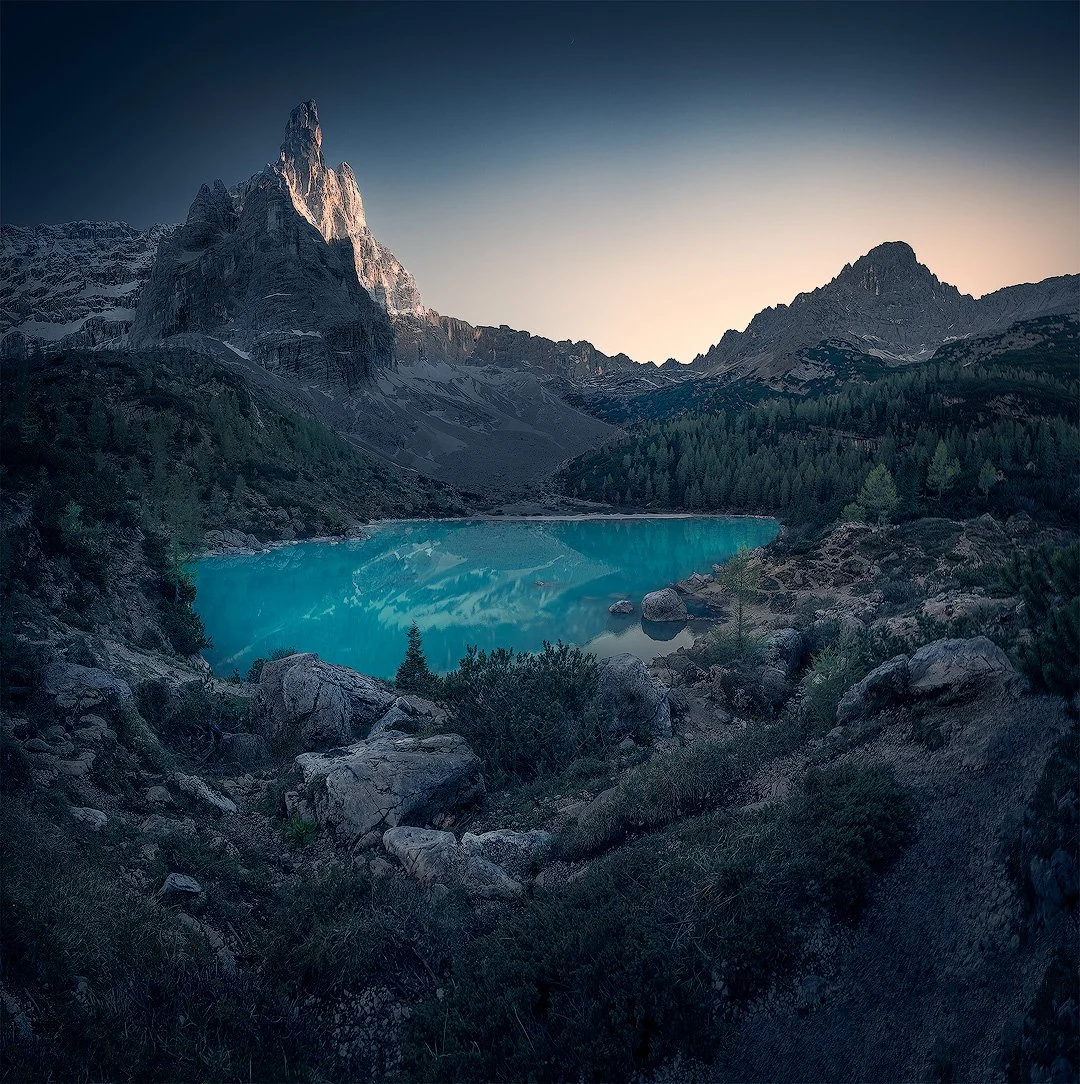 Bergsee mit türkisfarbenem Wasser, umgeben von Bergen und Wäldern, bei Sonnenaufgang oder Sonnenuntergang. Dolomiti.